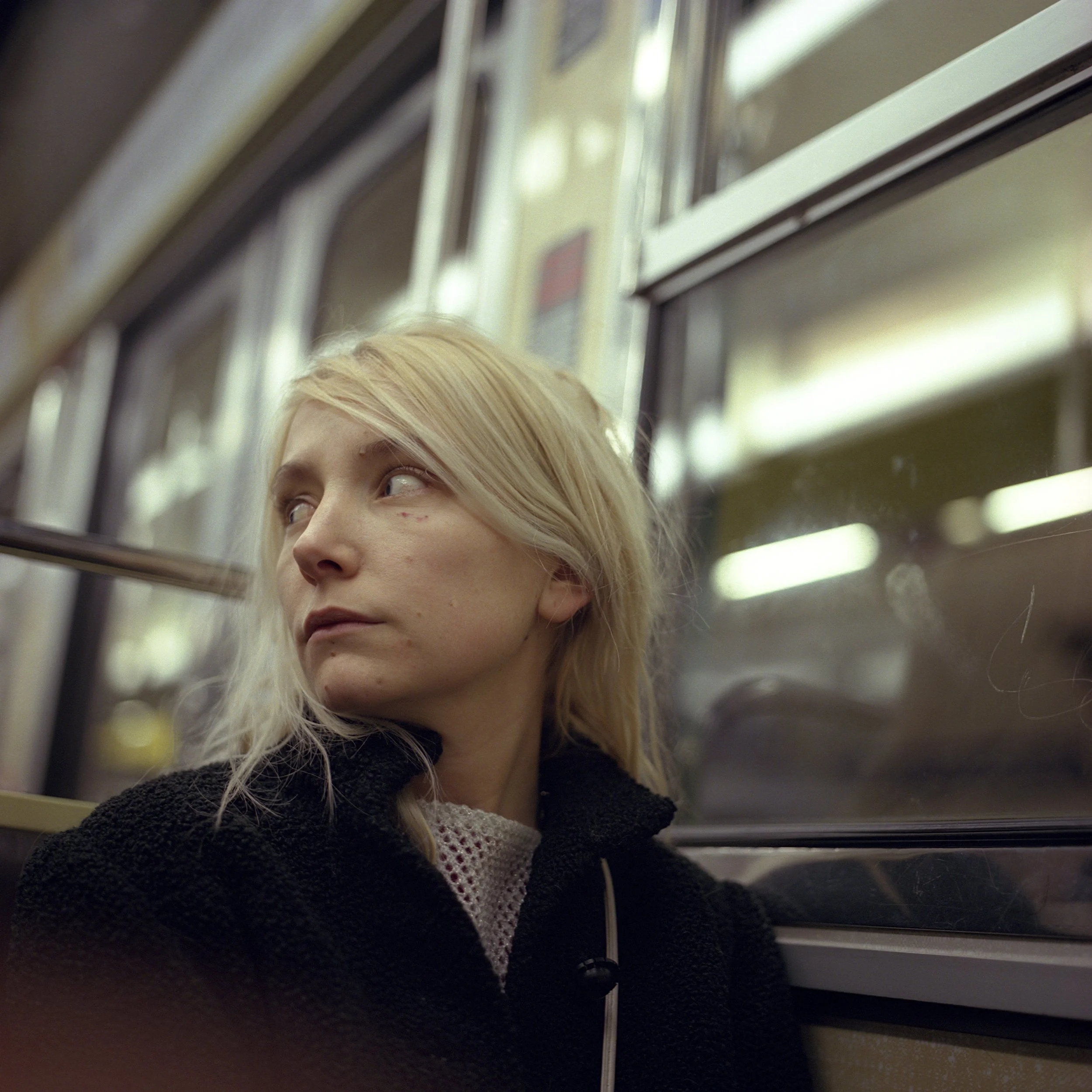A blonde woman with a black jacket sitting on a subway train, looking out of the window with a contemplative expression.