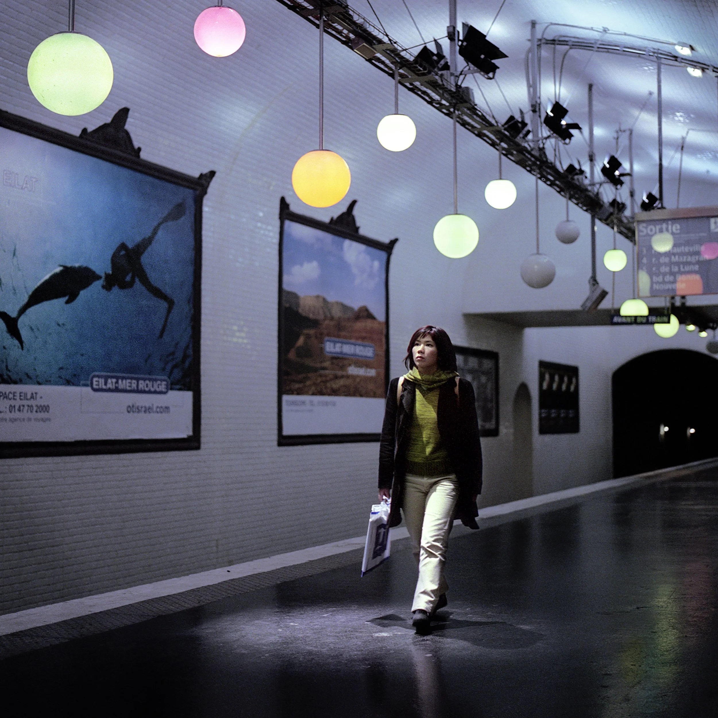 A woman walking through a dimly lit underground passage with colorful hanging lights and posters on the wall.