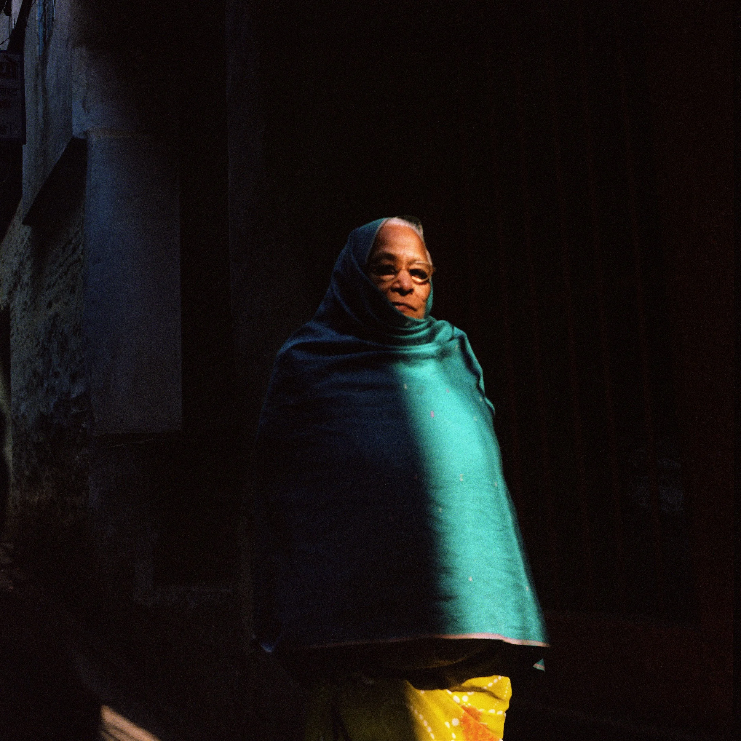 Older woman with glasses and grey hair wearing a teal shawl standing in a dimly lit space.