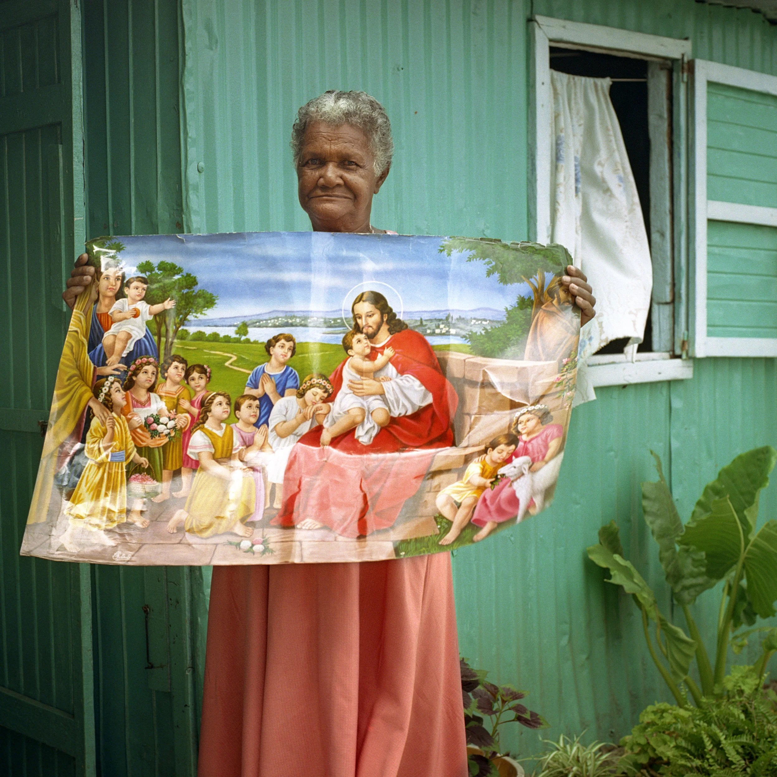 Mauritius. Trou d'Eau Douce. An elderly woman with short gray hair holding a religious poster in front of a green wooden house with an open window and cream curtains, surrounded by plants.