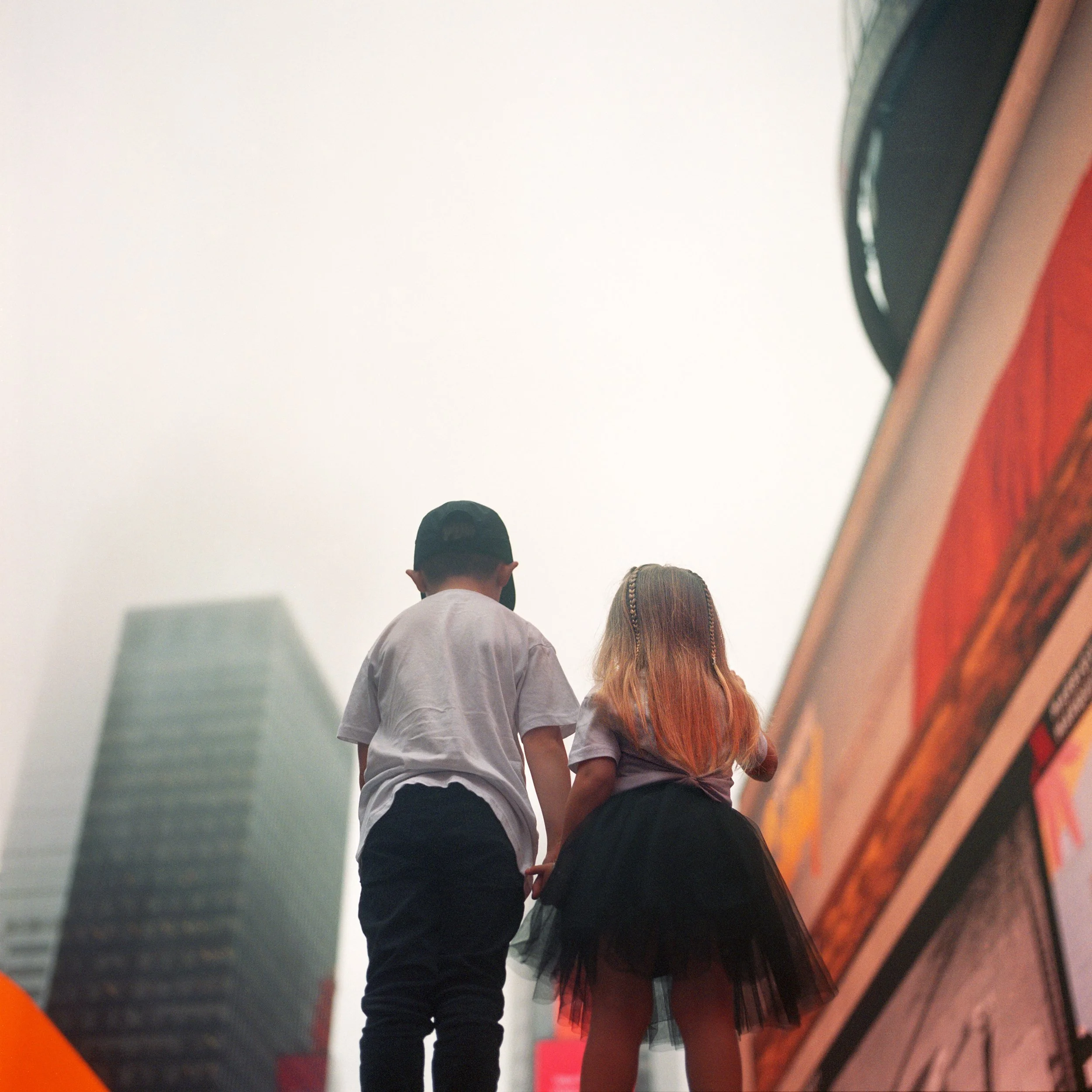 In New York Manhattan Times Sqaure, Two children, a boy and a girl, holding hands and looking upwards in a cityscape with tall buildings and a large screen or billboard.