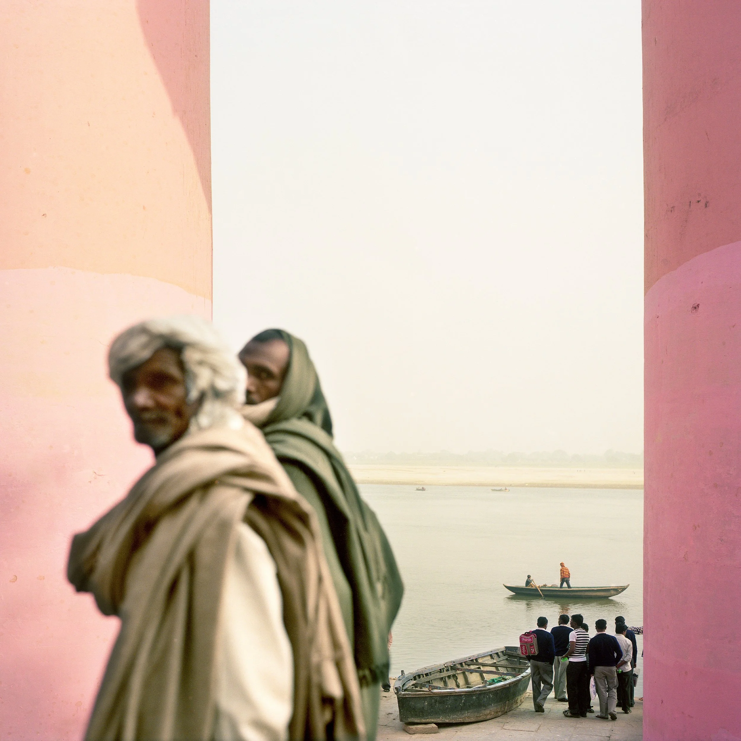 Two men in traditional clothing standing near an opening with a river, boats, and a group of people visible in the background.