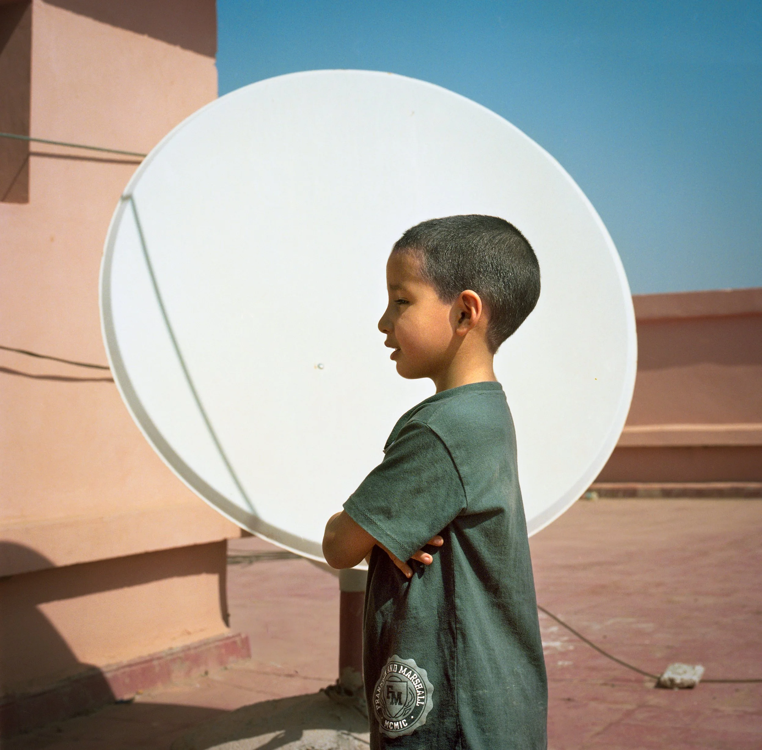 A young boy with short dark hair stands with arms crossed in front of a large satellite dish on a rooftop with pink and blue walls.