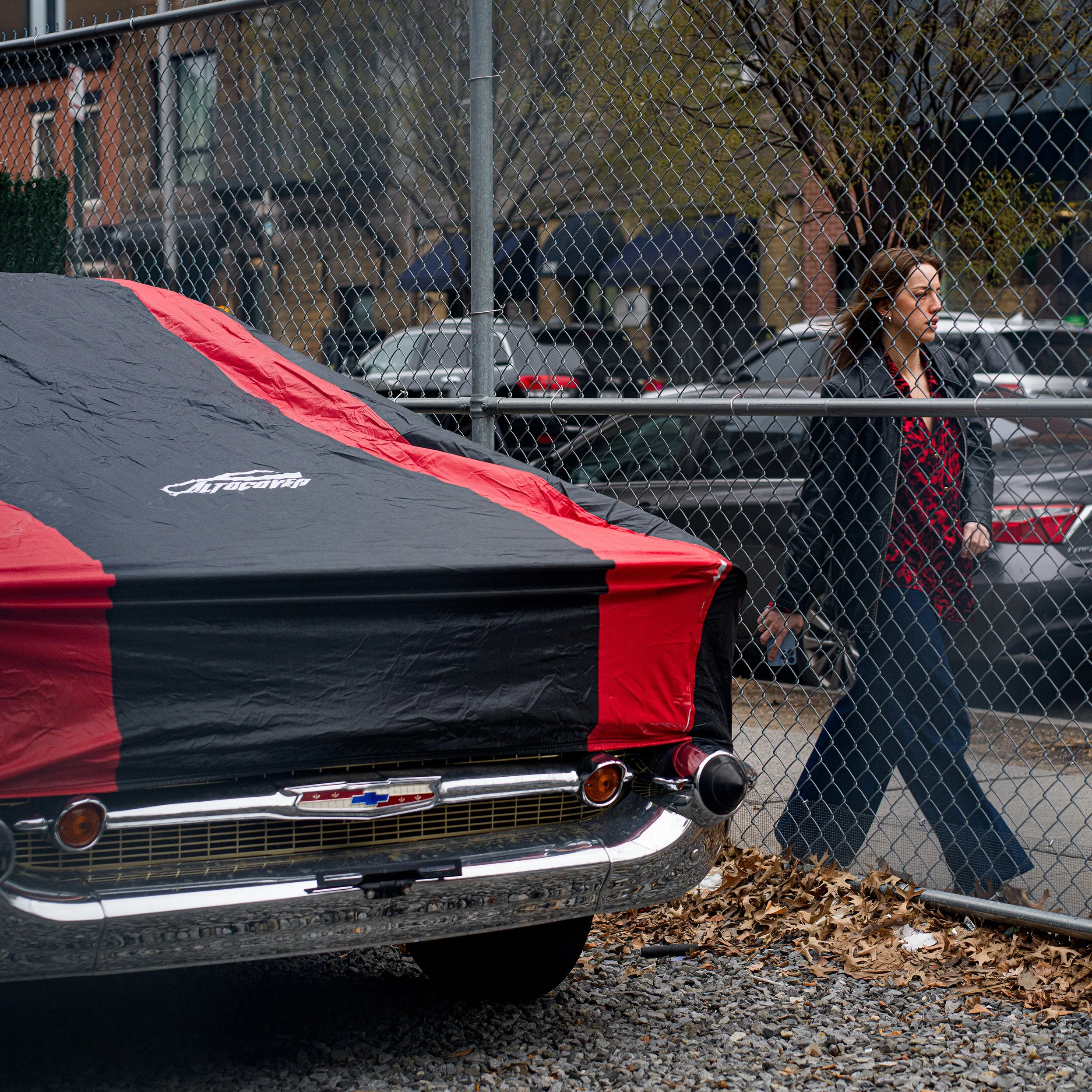	
In New York Manhattan, A woman walking past a chain-link fence, with a red and black covered vintage Chevrolet car parked nearby.