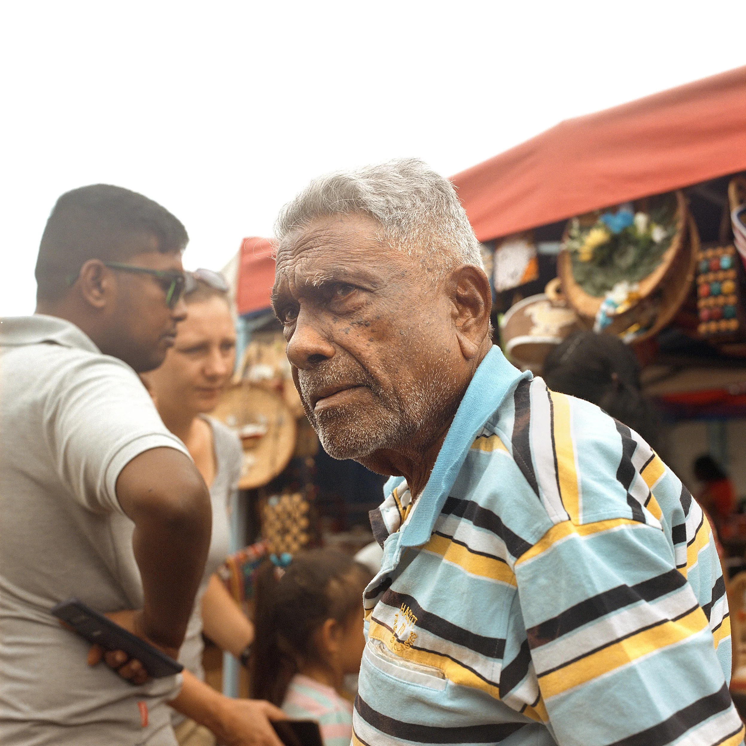 An elderly man with gray hair and a striped polo shirt at a market, with other people and colorful market stalls in the background.