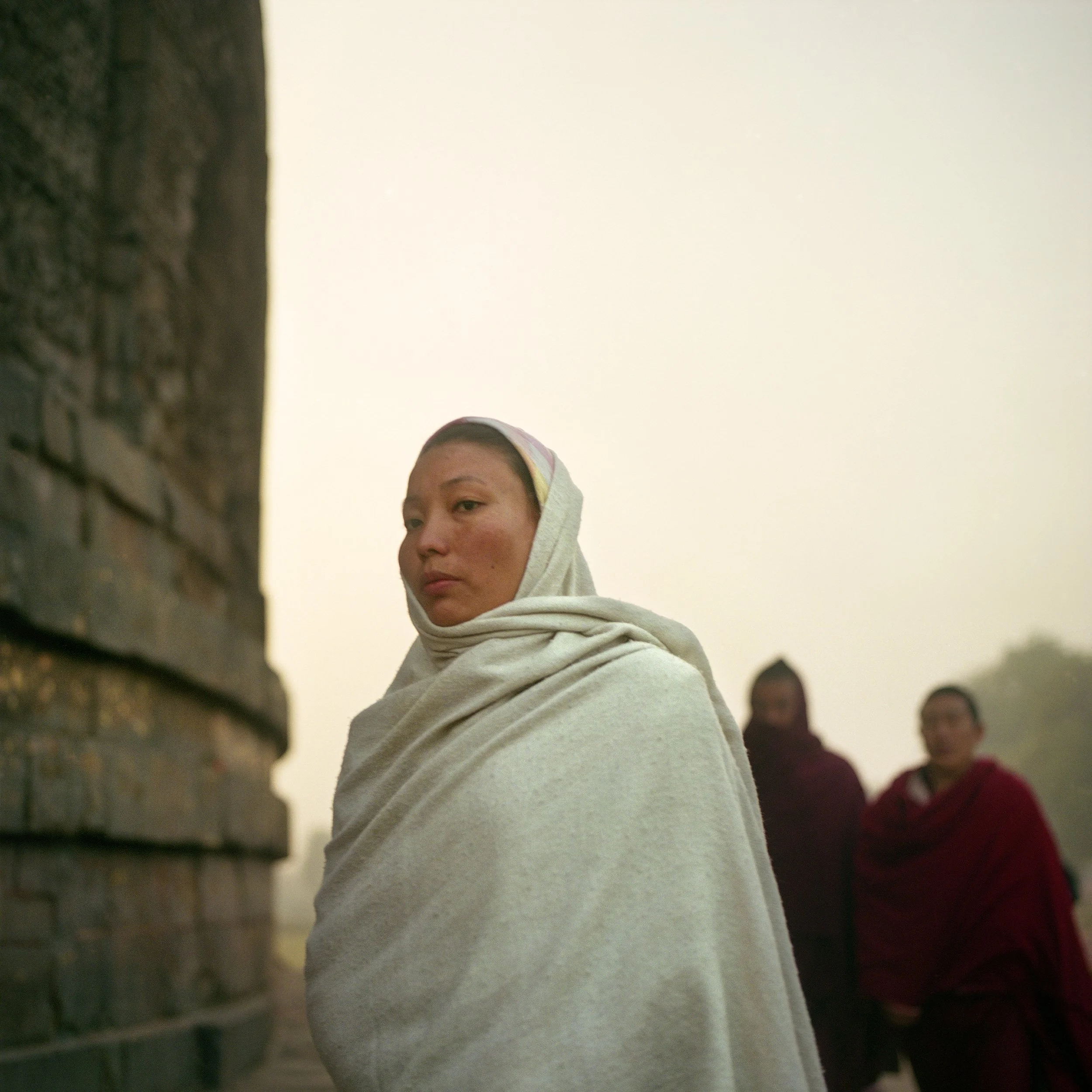 A woman in traditional attire wrapped in a white shawl with a head covering, standing outdoors with two men in the background, against a misty sky and distant landscape.