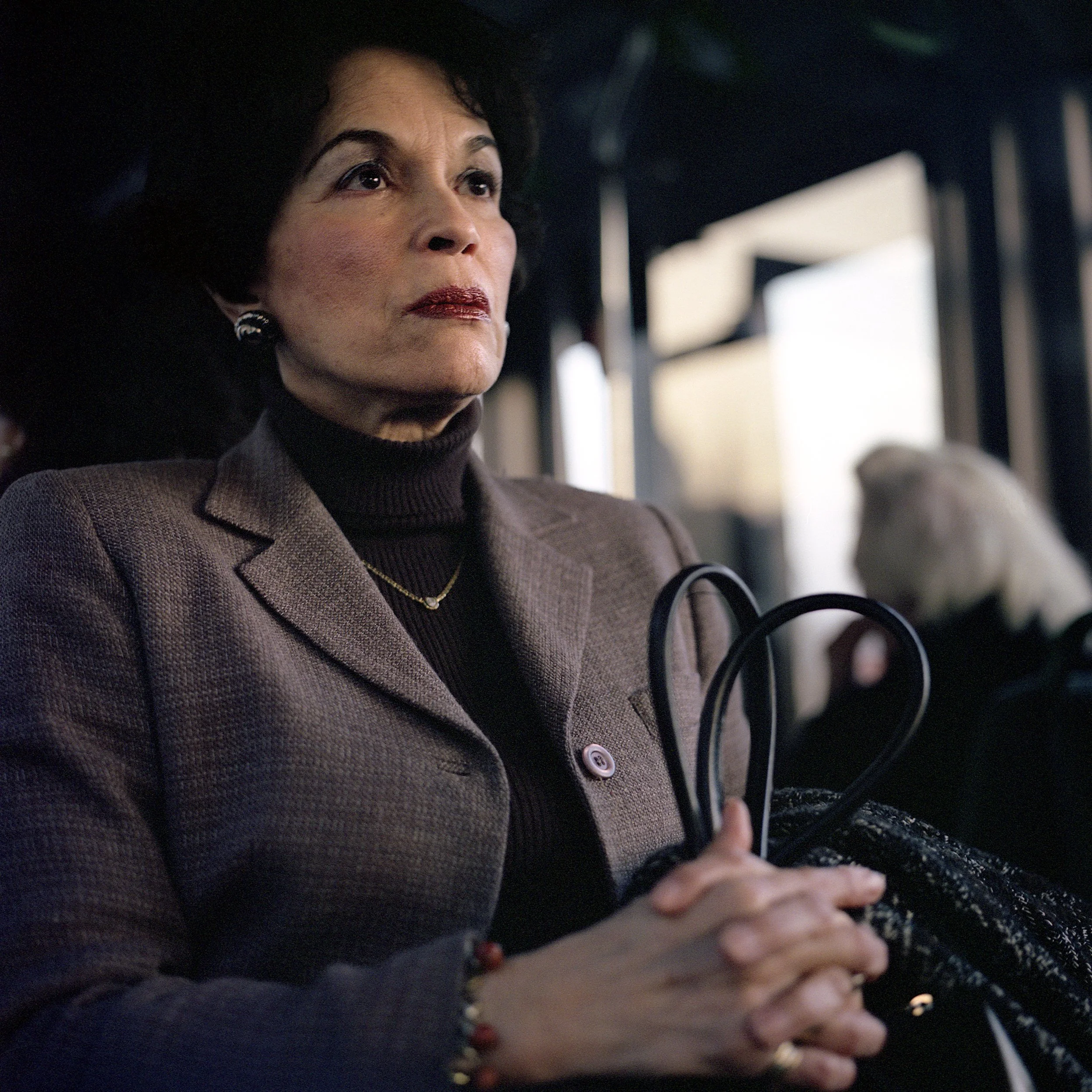 A woman with dark hair and wearing a brown blazer and turtleneck sits on a bus holding an umbrella. She has jewelry including earrings, a necklace, and bracelets. The background is blurred, showing a seated person with blonde hair.