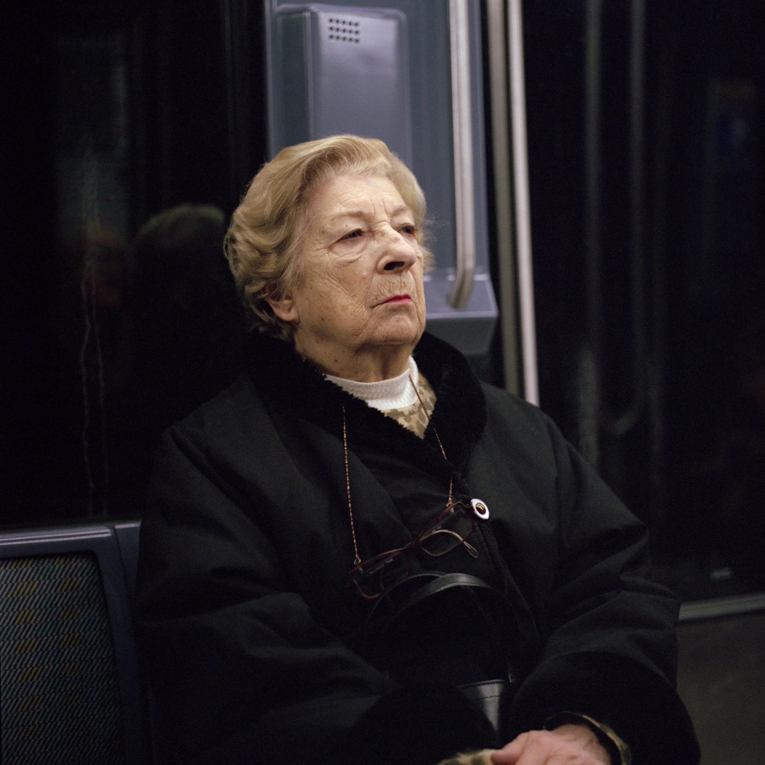 An elderly woman in Paris Subway with short, wavy blonde hair sitting with her arms crossed, wearing a black jacket and glasses hanging from her neck, in a subway train.
