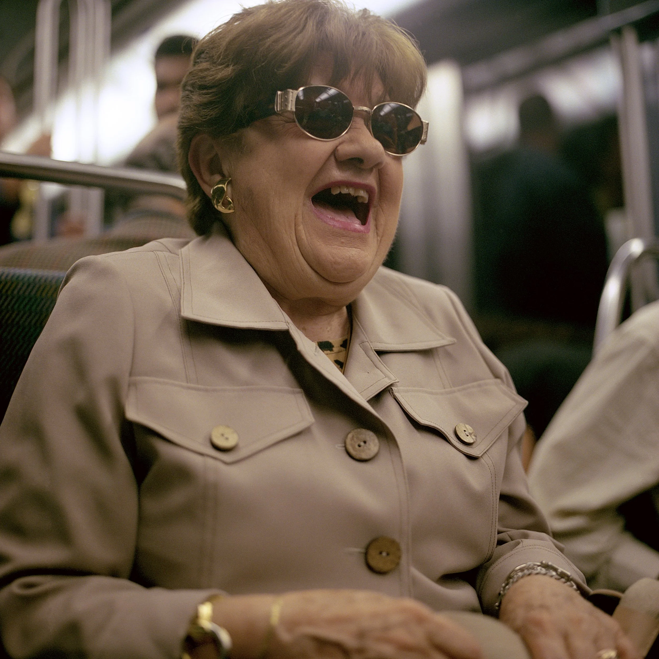 A cheerful elderly woman wearing sunglasses and a beige trench coat, smiling and laughing while sitting on a bus.