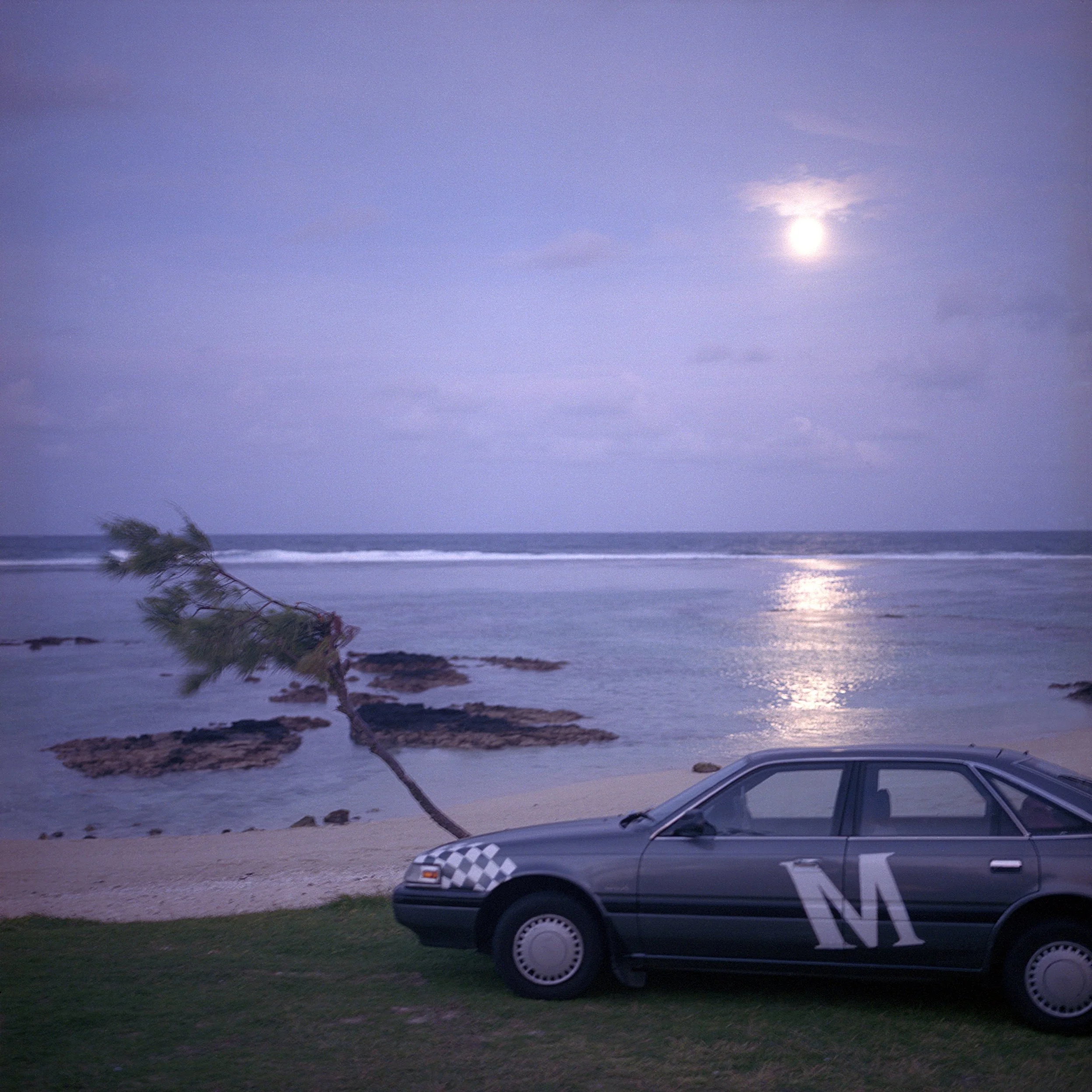 IN Palmar, Trou d'eau douce, Mauritius, A car parked on a grassy area by the beach with a fallen filao tree and a large moon in the sky.