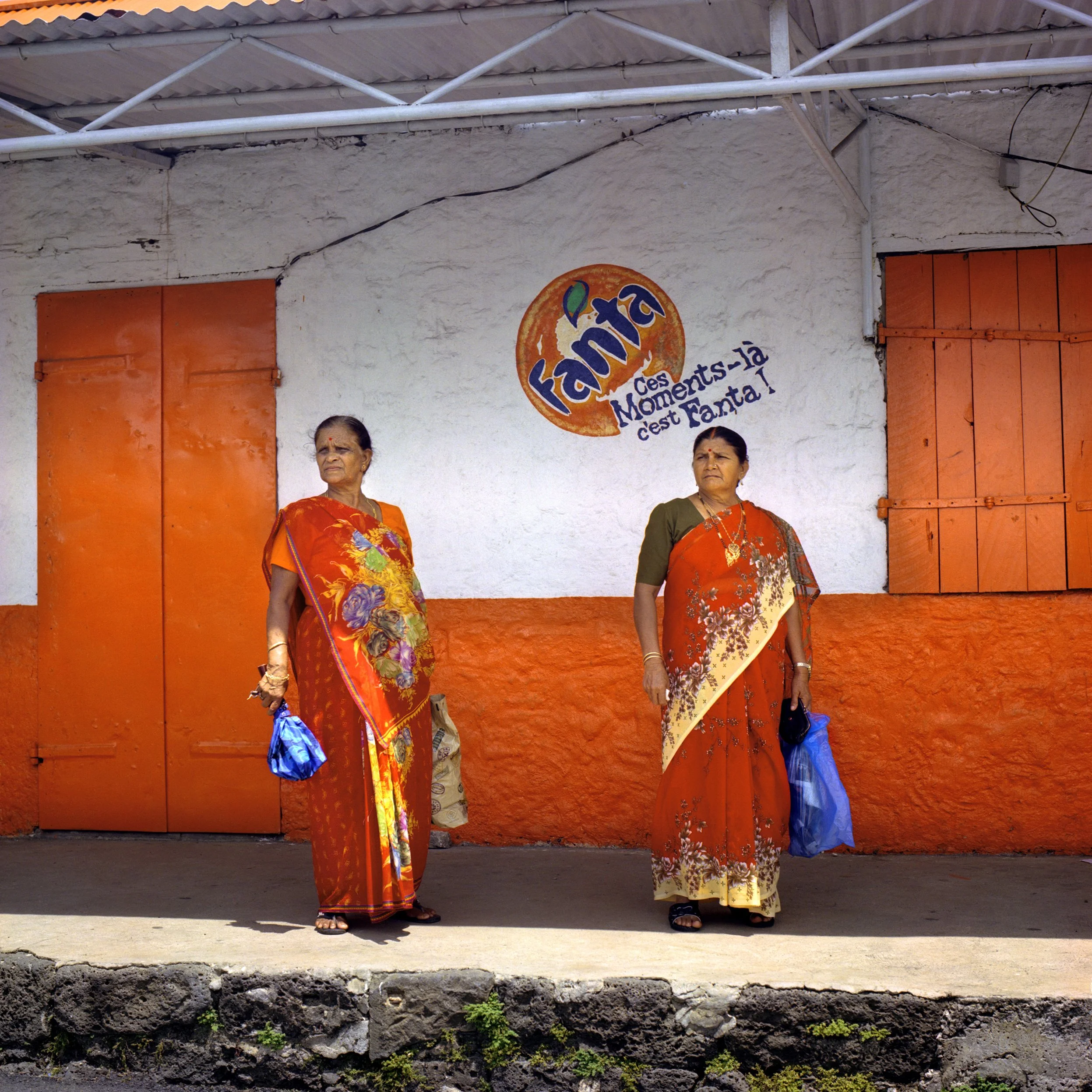 In Mauritius, Trou d'Eau Douce, Two women dressed in traditional Indian sarees standing on a sidewalk in front of a wall painted orange and white. The wall features a logo for Fanta and the French slogan Ces moments là c'est Fanta