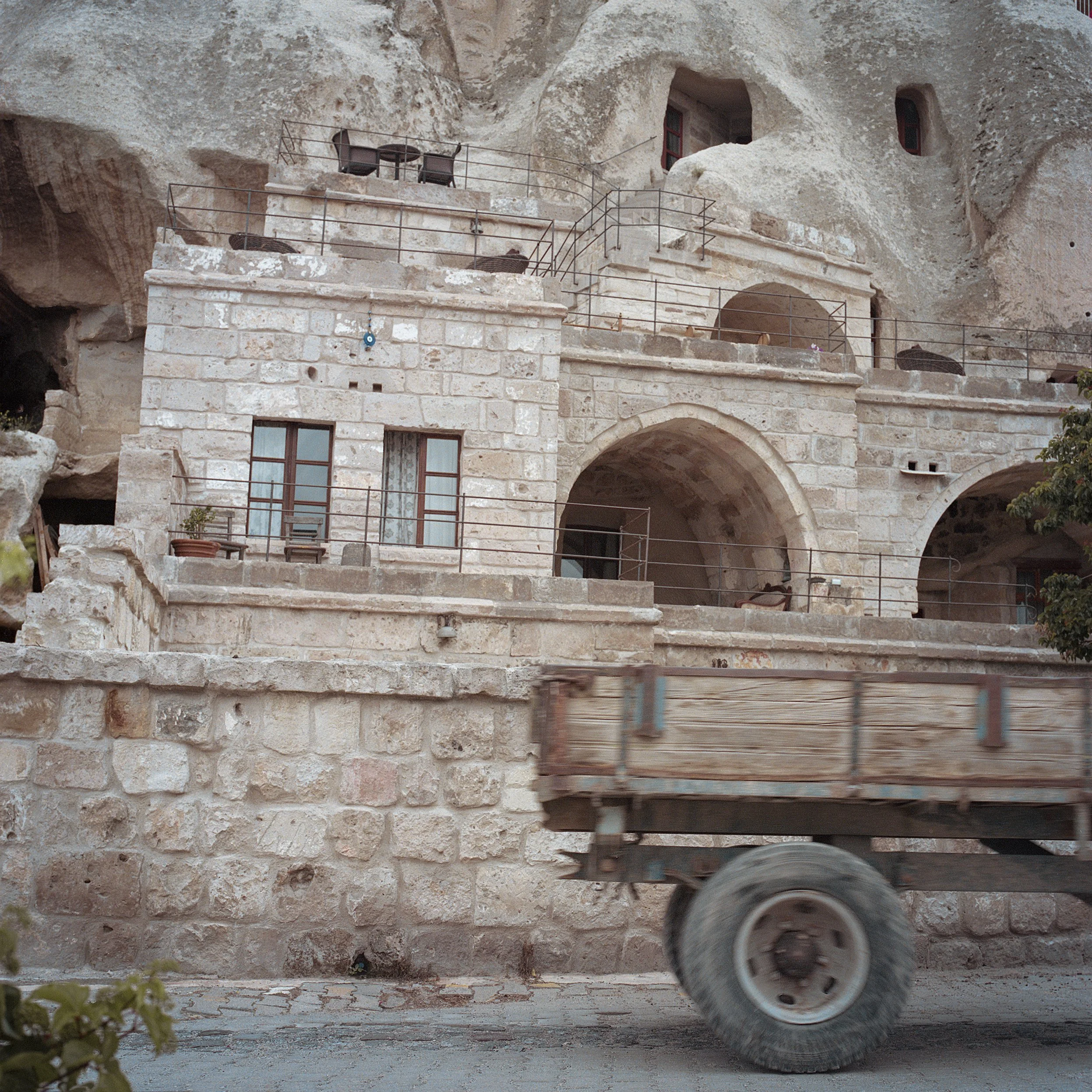In Goreme, Turkey, Stone building built into a cave with multiple levels, arches, and windows, with a moving truck in the foreground.