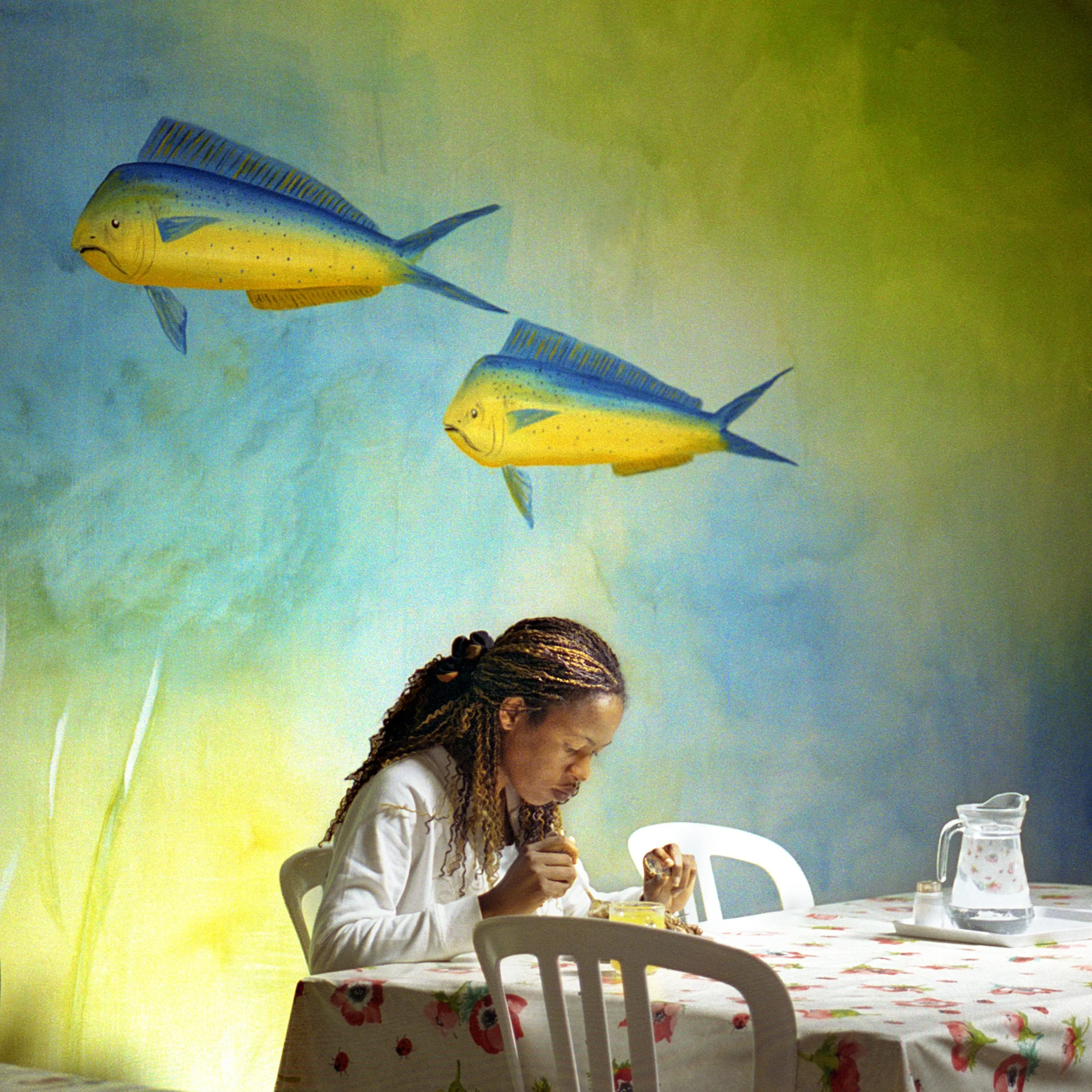 In Mauritius, A woman with braids is sitting at a table, eating, with a pitcher and a small bowl of food on the table. The background features two large, colorful fish swimming in an animated, surreal scene.