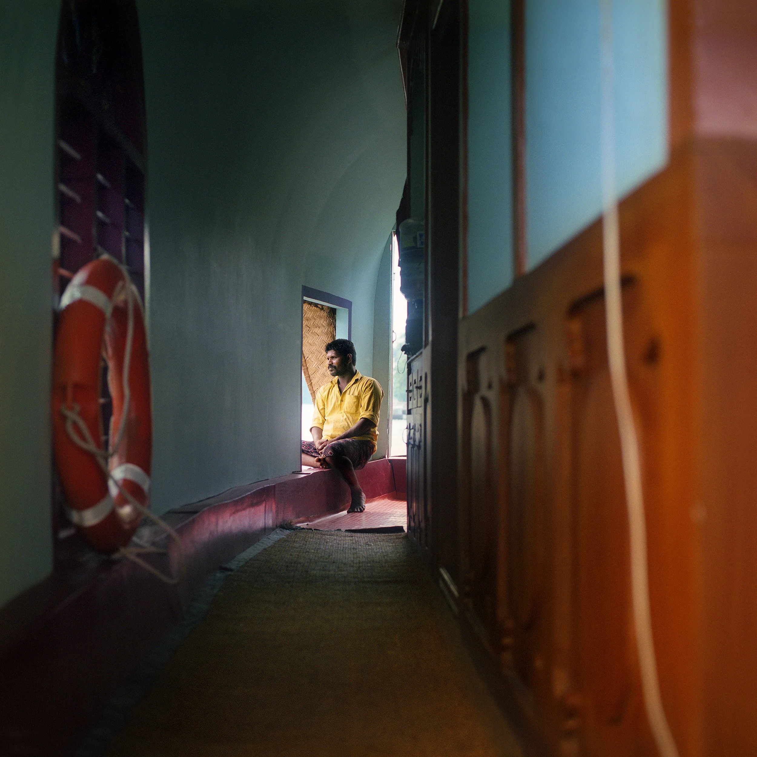 In Kerala backwaters, A man sitting in a boat, cross-legged on a window ledge, viewed from a narrow gap between furniture, with green walls and some framed pictures in the background.