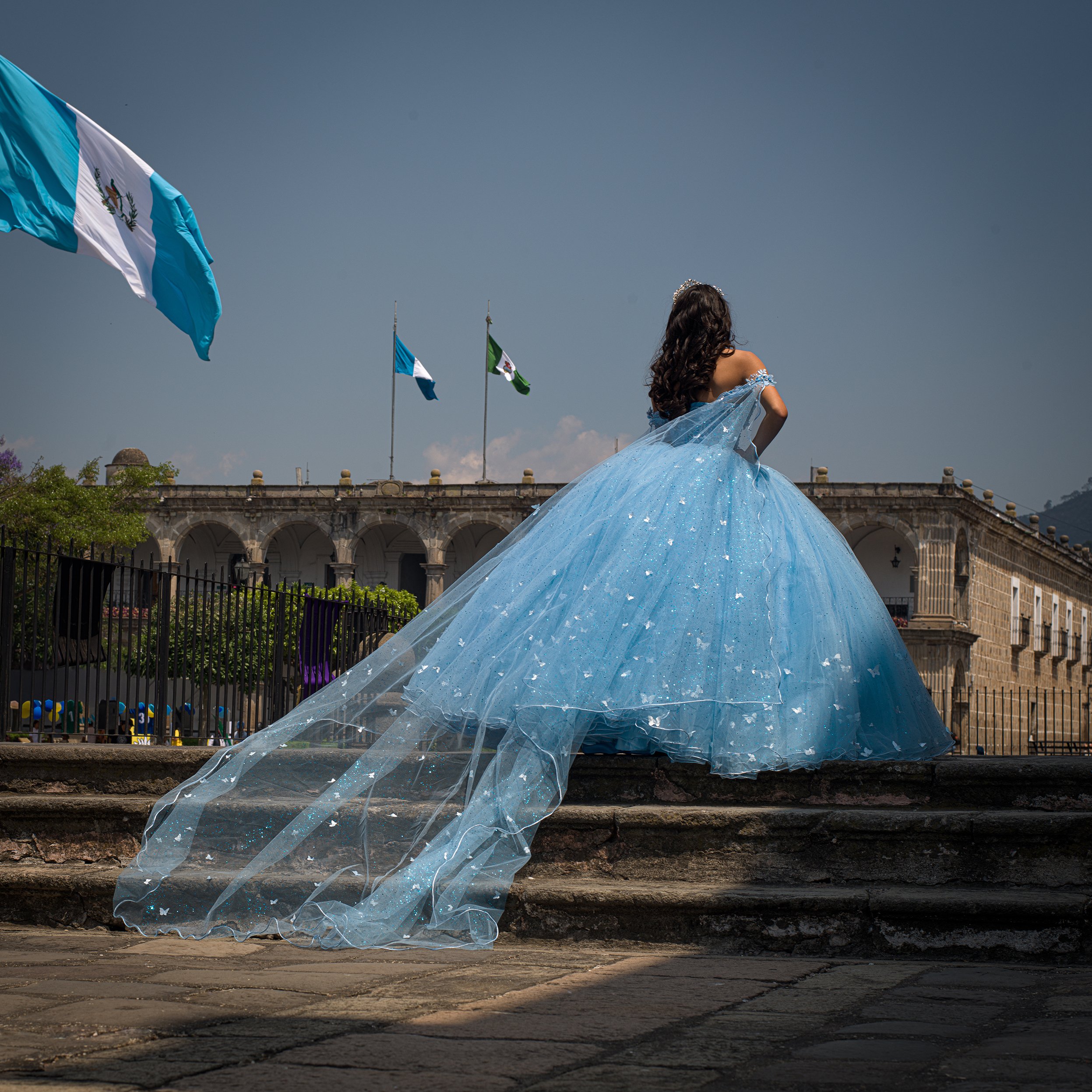 In Antigua, Guatemala, Quinceañera, A a5 years old girl dressed as a princess in a blue ball gown with a long, flowing train standing on stone steps in front of a historic building with flags, under a blue sky.