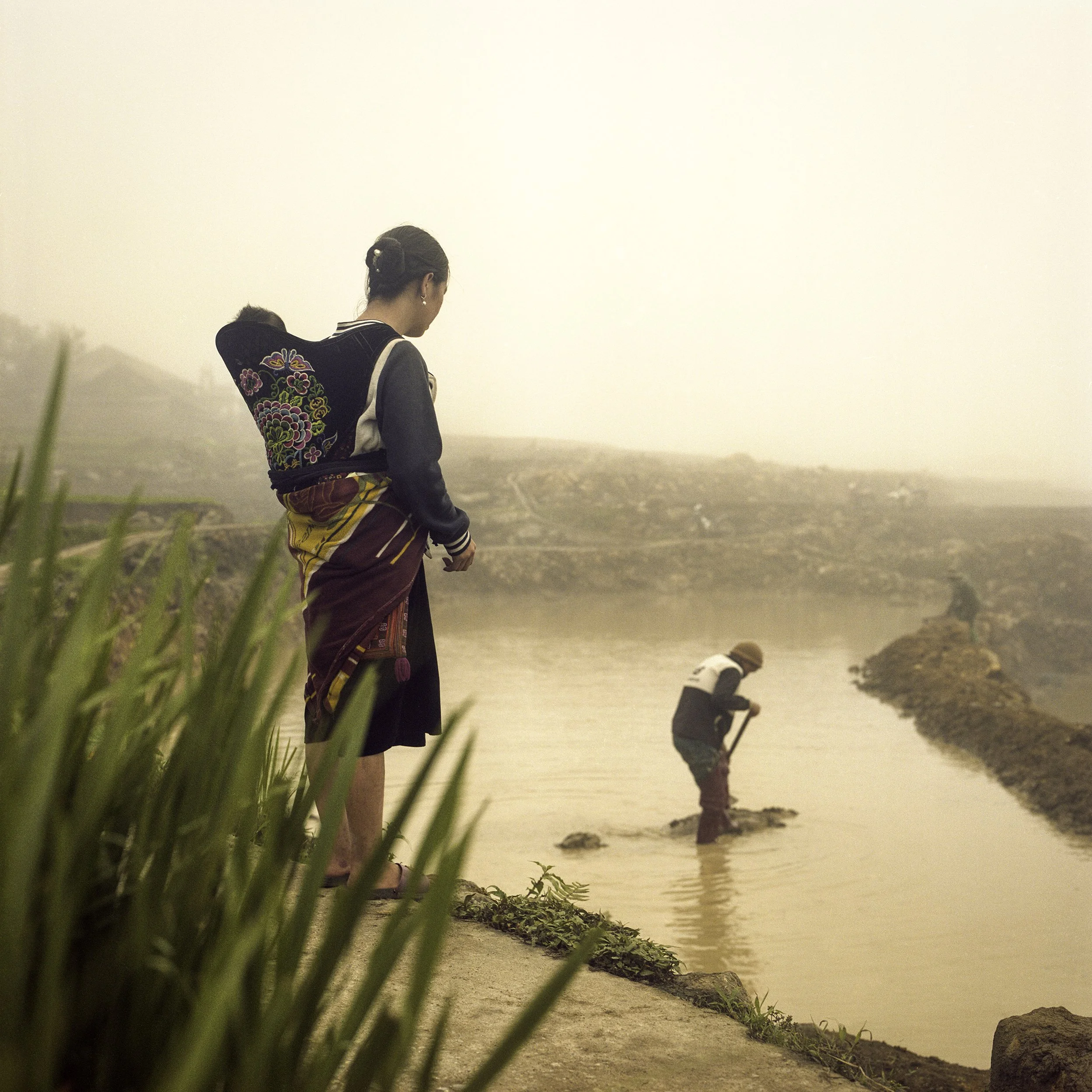 Two women stand near a riverbank on a foggy day, one standing on the shore and the other wading in the water, with misty hills in the background.