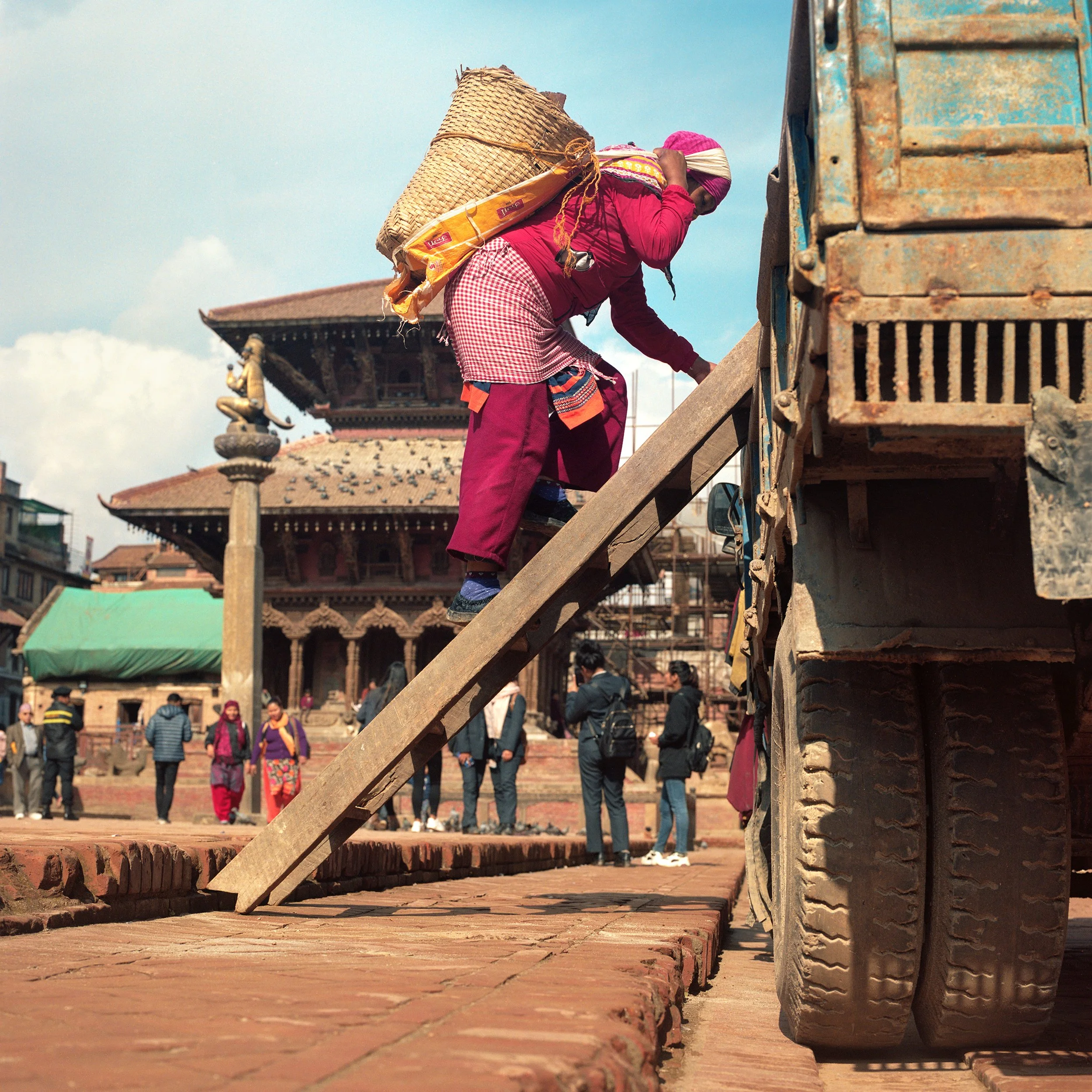 In Katmandu, A woman wearing pink clothing and a headscarf climbs a wooden ramp to a truck, with a historic building in the background and other people walking around.