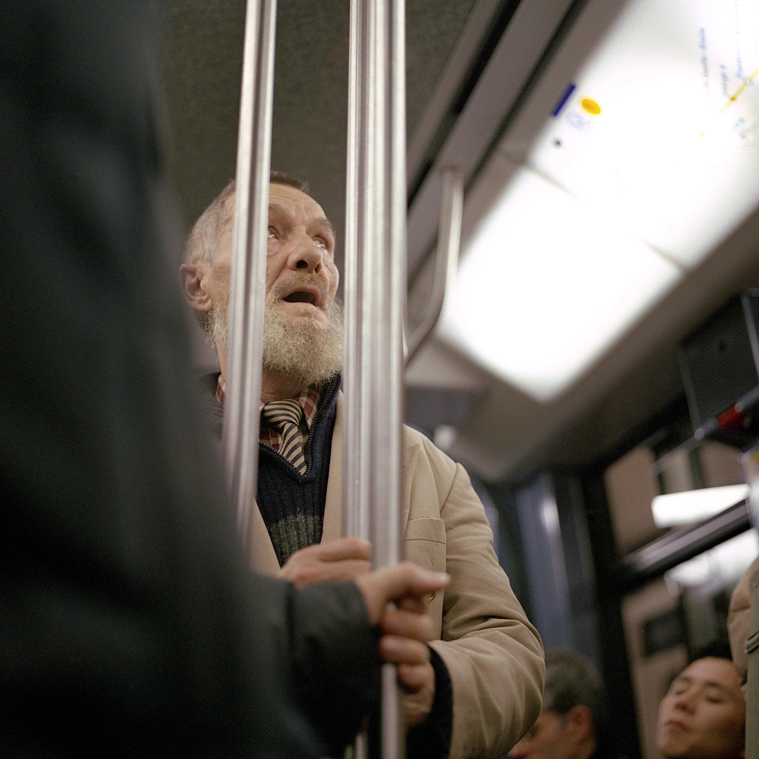 An elderly man with a beard holding onto vertical bars inside a train or subway car, looking upwards with a distressed or scared expression.