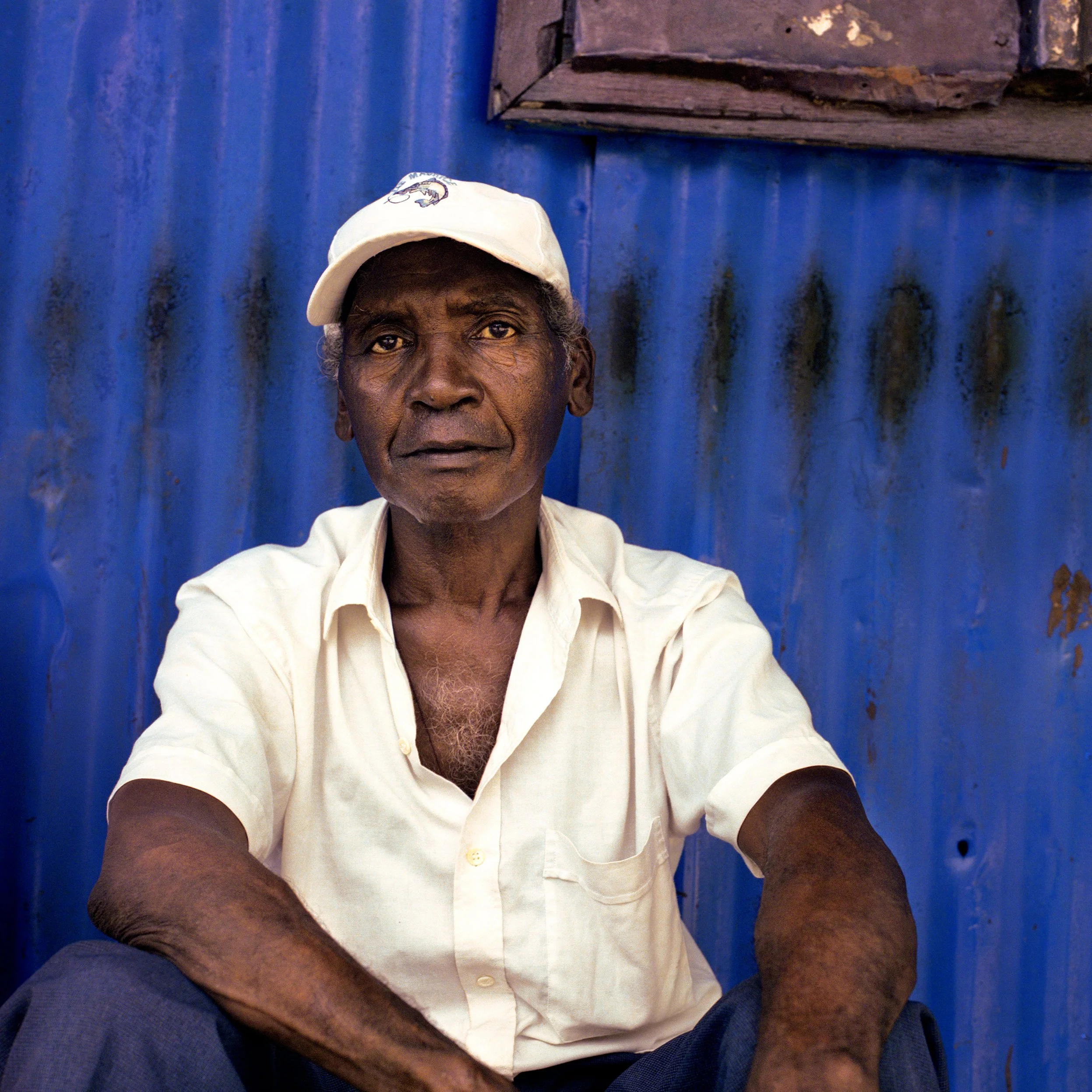 In Mauritius, Trou d'Eau Douce, An older man sitting against a blue corrugated metal wall, wearing a white shirt and a white cap.