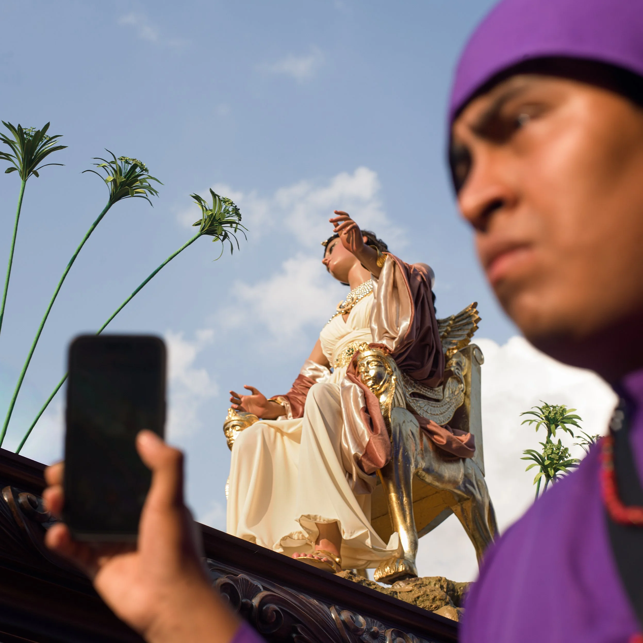 In Antigua, Guatemala, Alfombras, A man wearing a purple head covering takes a selfie with a large golden statue of a seated woman in the background, outdoors with a partly cloudy sky, tall plants, and decorative elements.
