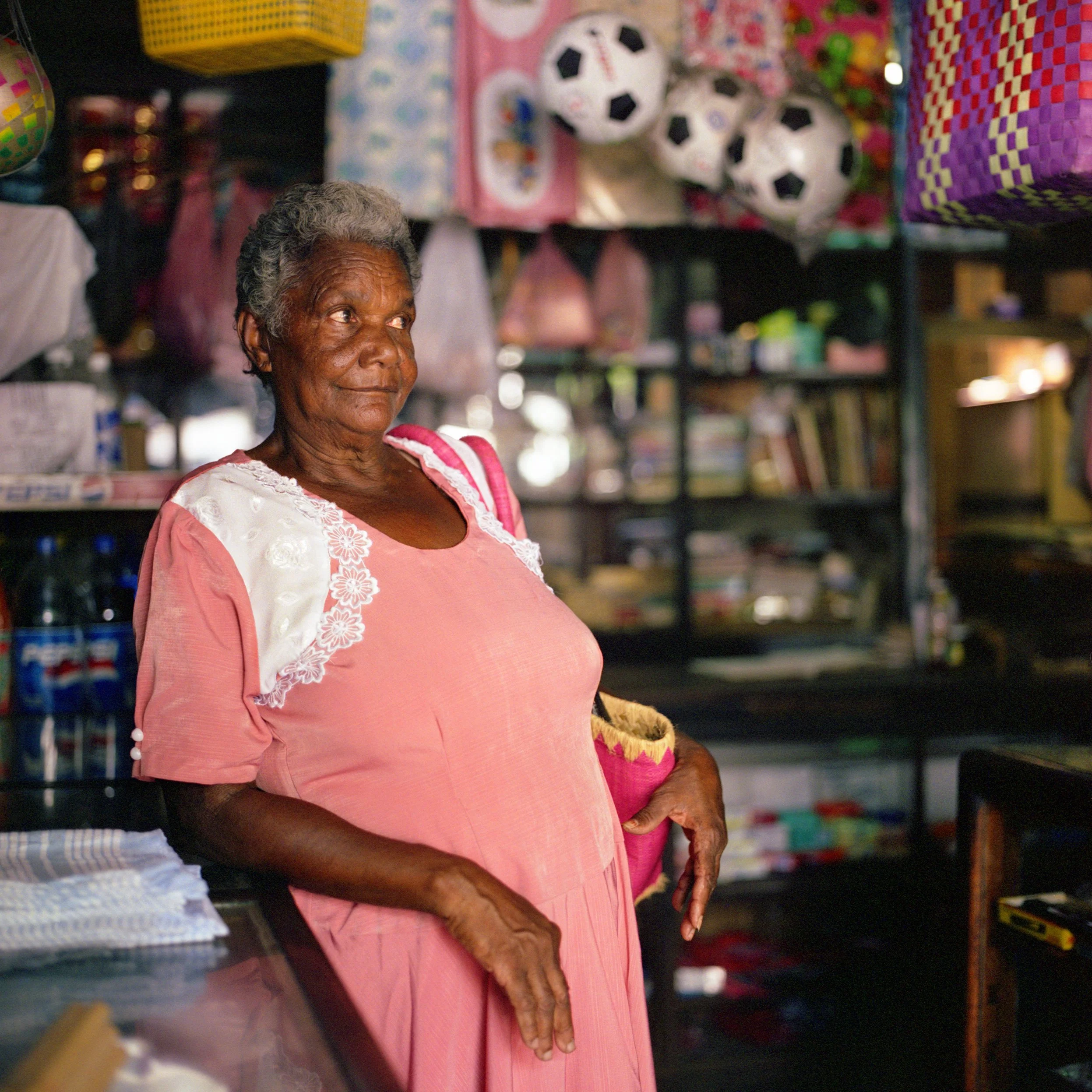	
In Mauritius, Flack, An elderly woman with short gray hair wearing a pink dress with white lace details, standing inside a store with shelves filled with toys, books, and balloons around her.