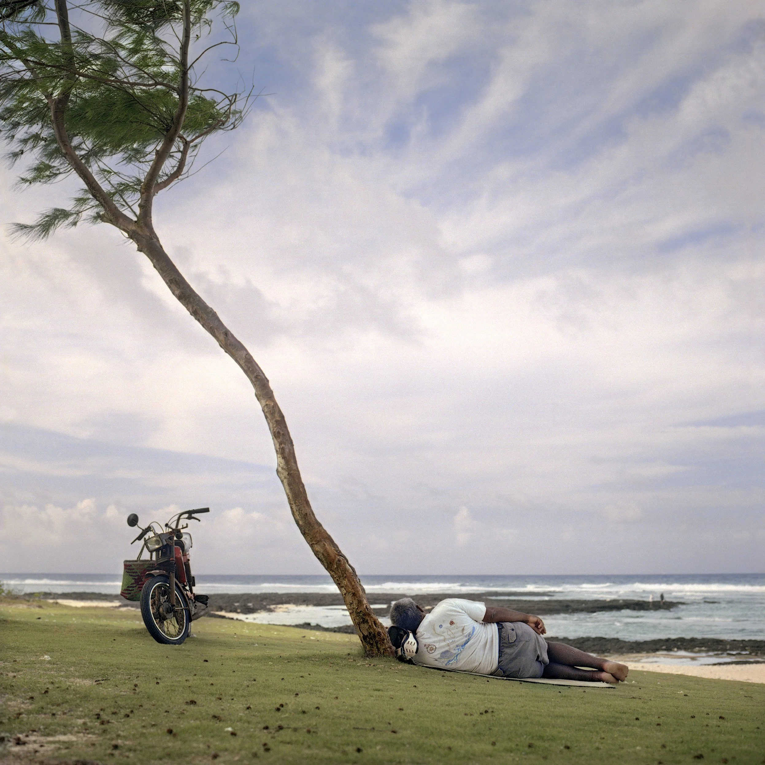 In Mauritius, Palmar public beach, A man lying on the grass near a shoreline with a bicycle and a tree beside him, with a cloudy sky overhead.