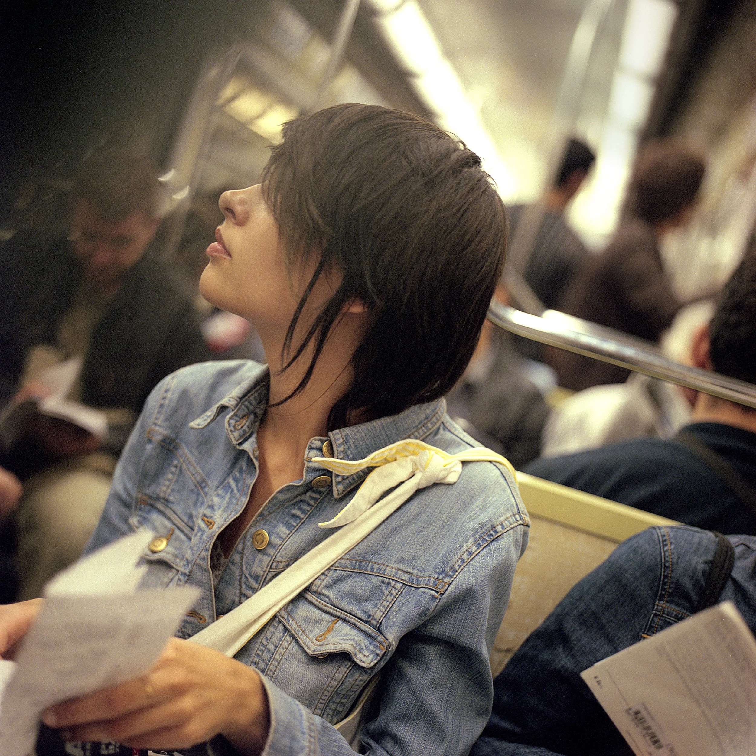A woman with shoulder-length dark hair on a subway train, sitting and looking to her left, wearing a denim jacket and holding a piece of paper.
