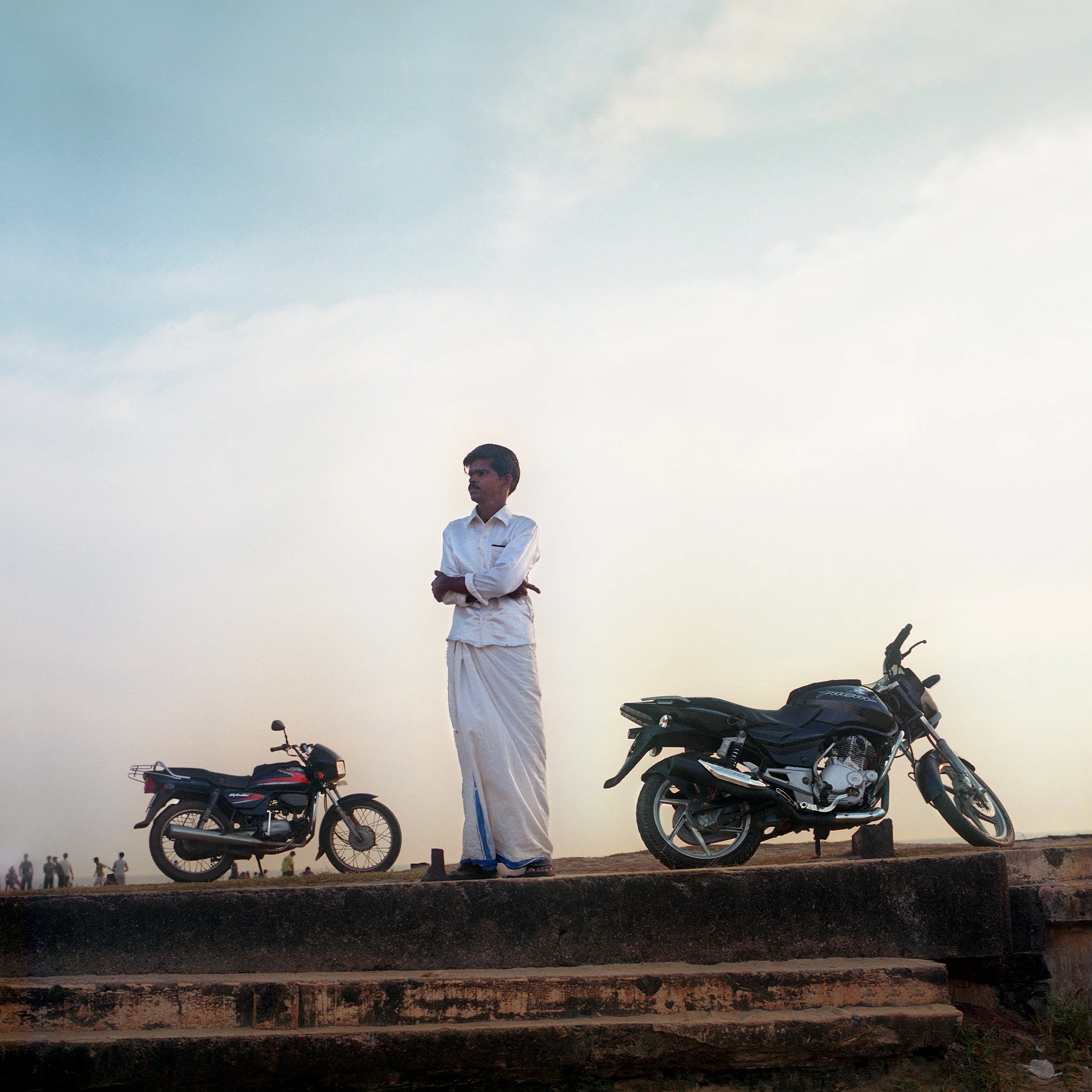 In Kerala, India, A man dressed in traditional Indian attire standing on stone steps outdoors, with two motorcycles parked nearby, under a cloudy sky.