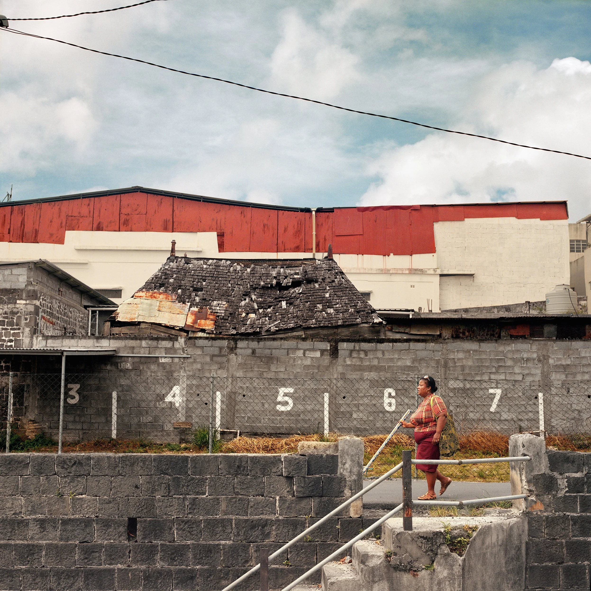 A woman walking along a sidewalk in front of a gray brick wall with white painted numbers 3 to 7, behind which is an old building with a damaged roof, and a background of modern buildings with a red roof and a cloudy sky.