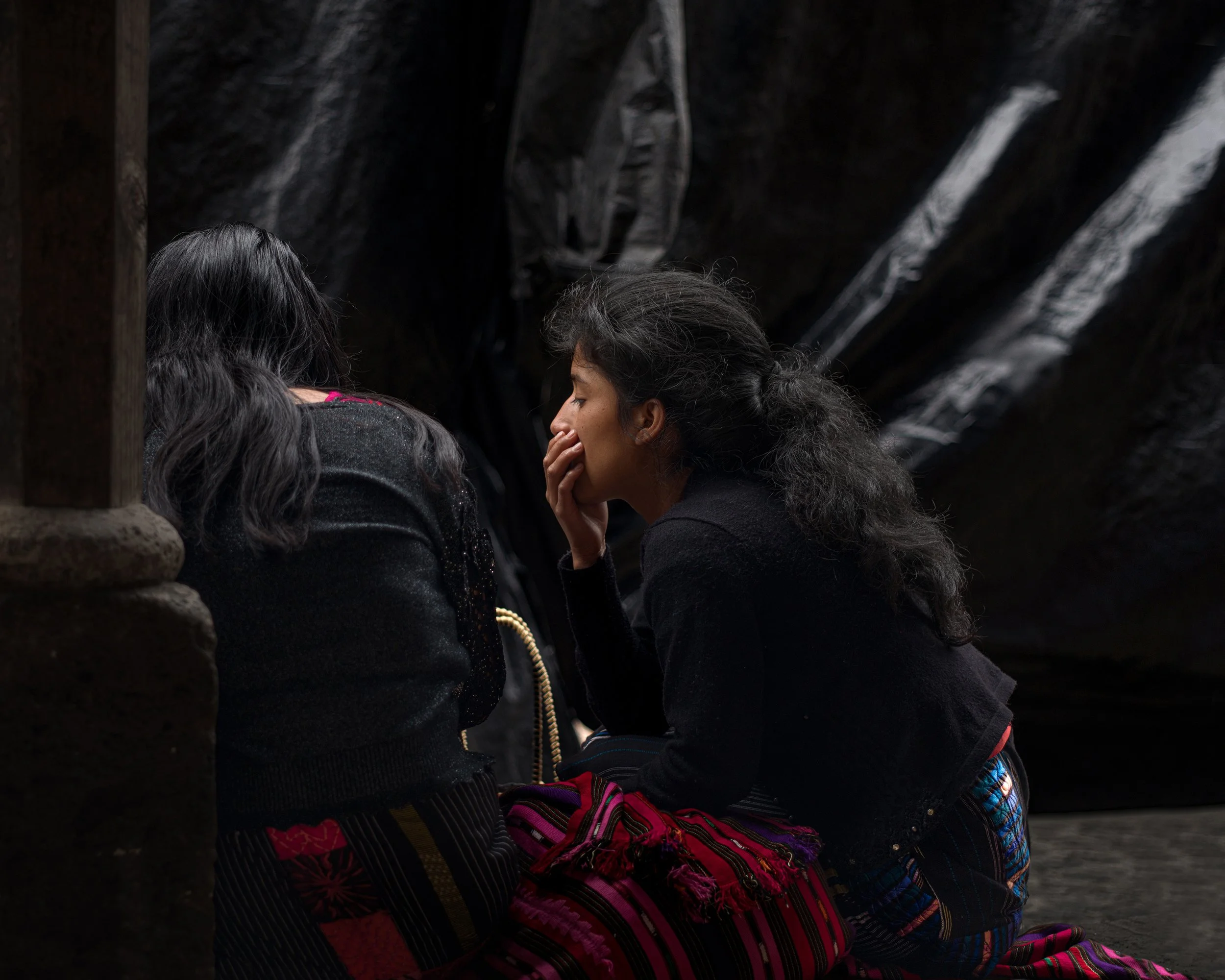 In Chichicastenango market. Guatemala, Two women sitting close together in an indoor or cave-like setting, engaging in a private and serious conversation, with one woman covering her mouth with her hand.