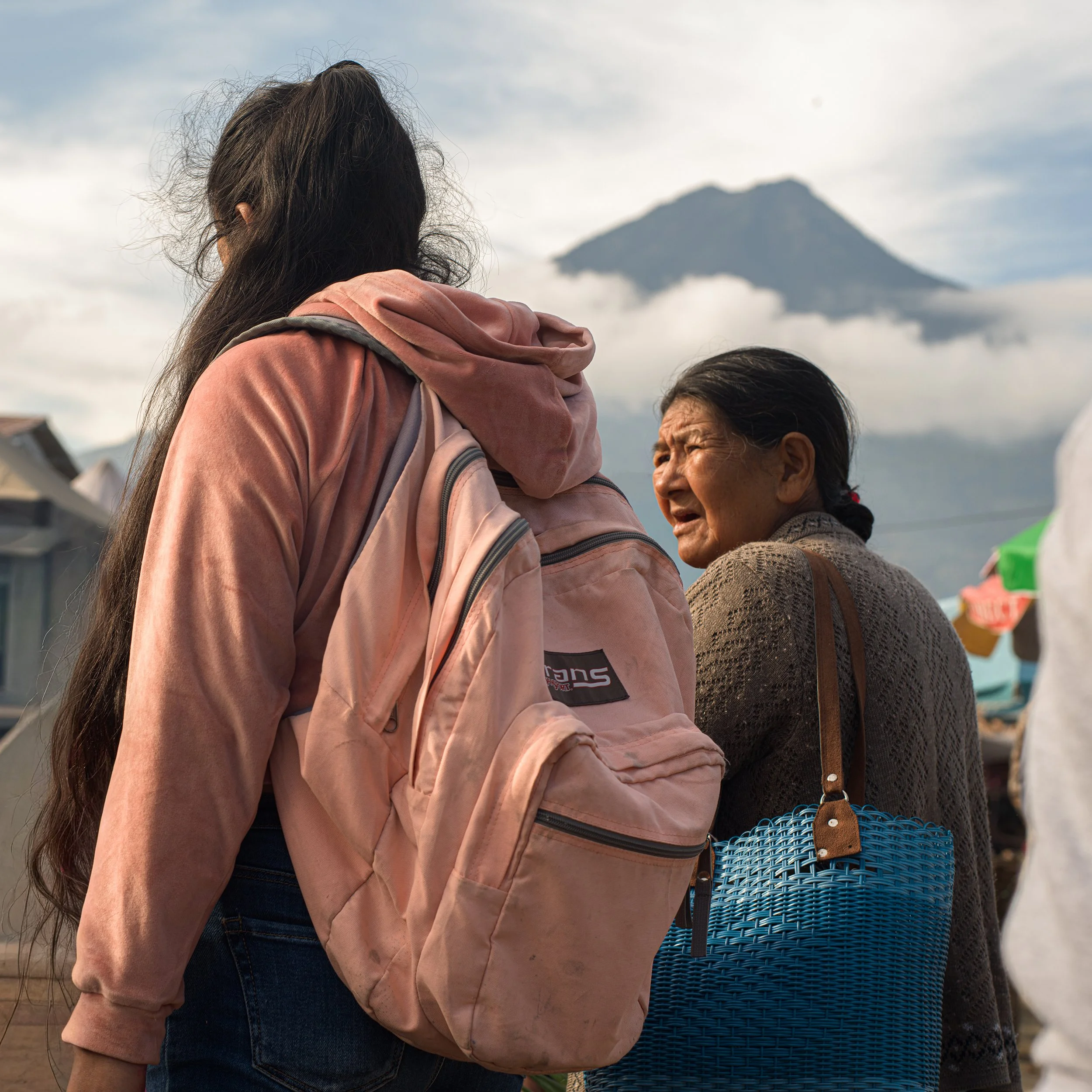 	
In Antigua market, Guatemala, A young woman with a pink backpack talking to an elderly woman with a blue woven basket in a marketplace with a mountain in the background.
