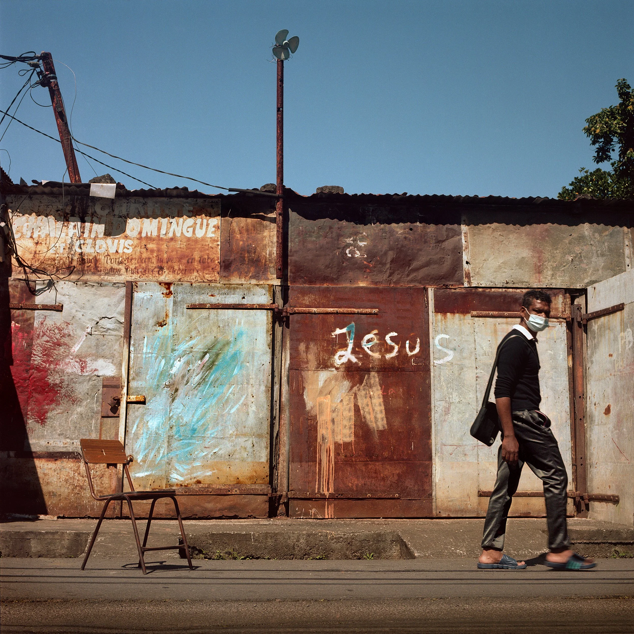 A man walking past a rusty metal wall with the word 'Jesus' spray painted on it, wearing a face mask, black sweater, and gray pants, with a shopping bag on his shoulder; an empty chair on the sidewalk in front of him.