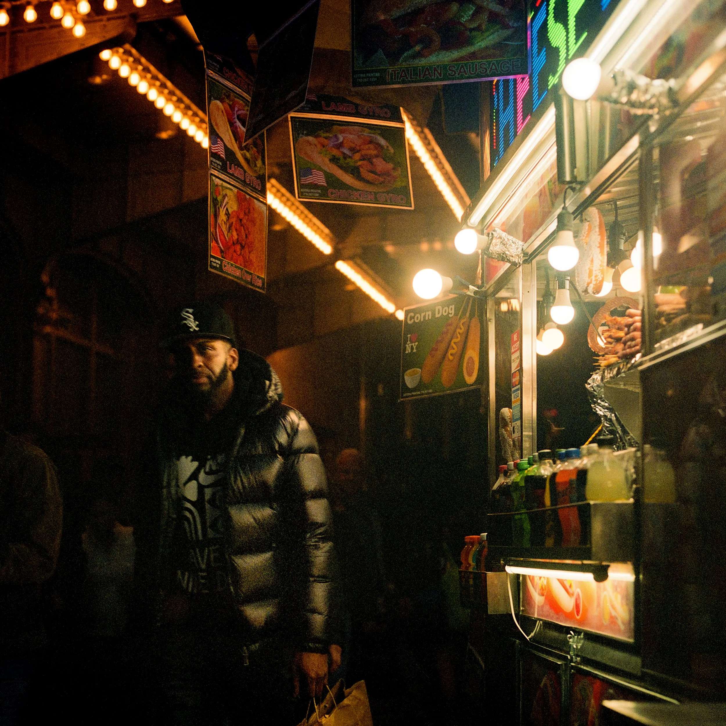 In New York Manhattan, A man in a black jacket and baseball cap shopping at a food stand illuminated by bright lights in a nighttime street scene.