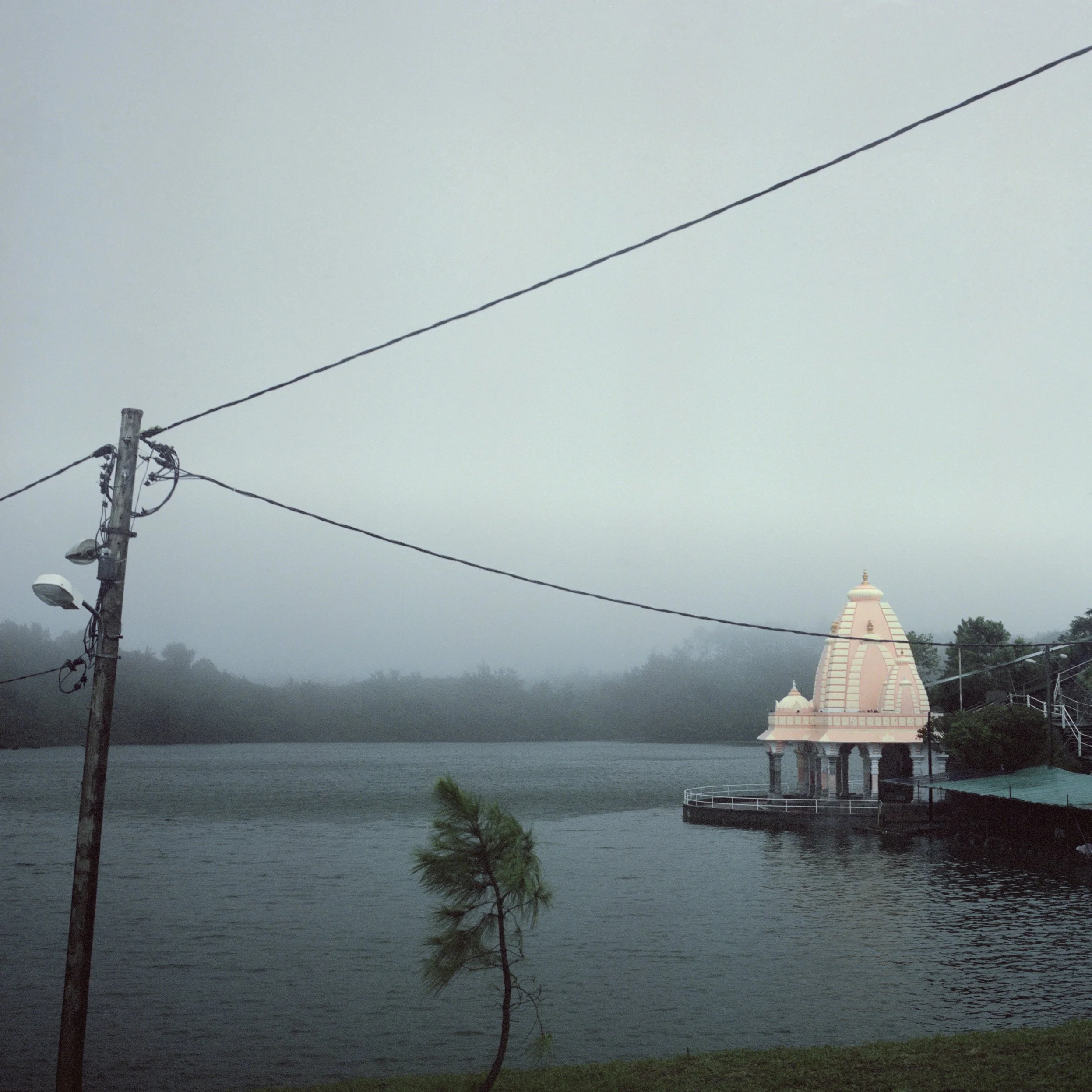In Mauritius, Ganga Talao, A stormy scene with a foggy lake, a utility pole with power lines, and a temple in the background.