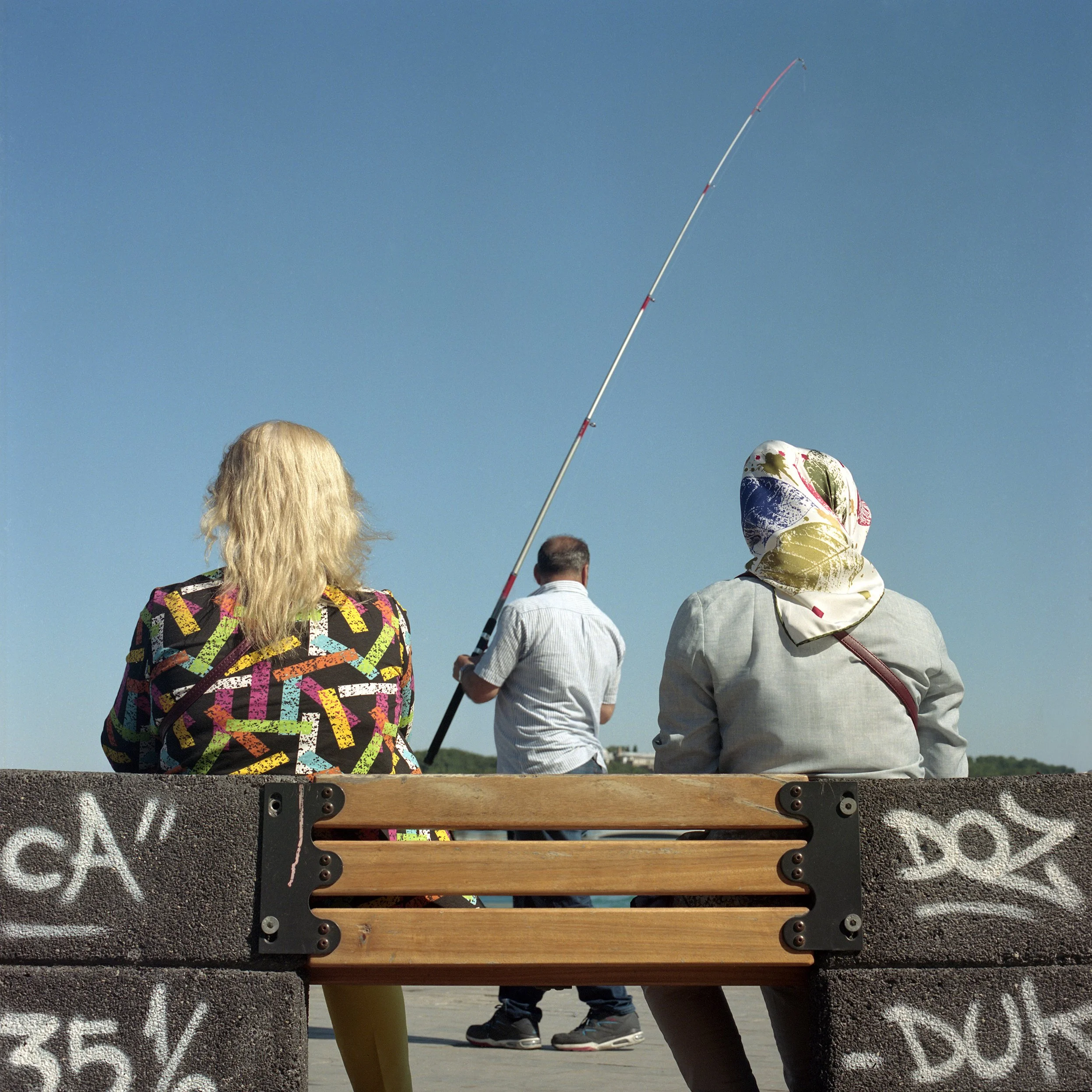 In Istanbul, Turkey, Three people sitting on a bench by the water, watching a man fishing with a tall fishing pole under a clear blue sky.