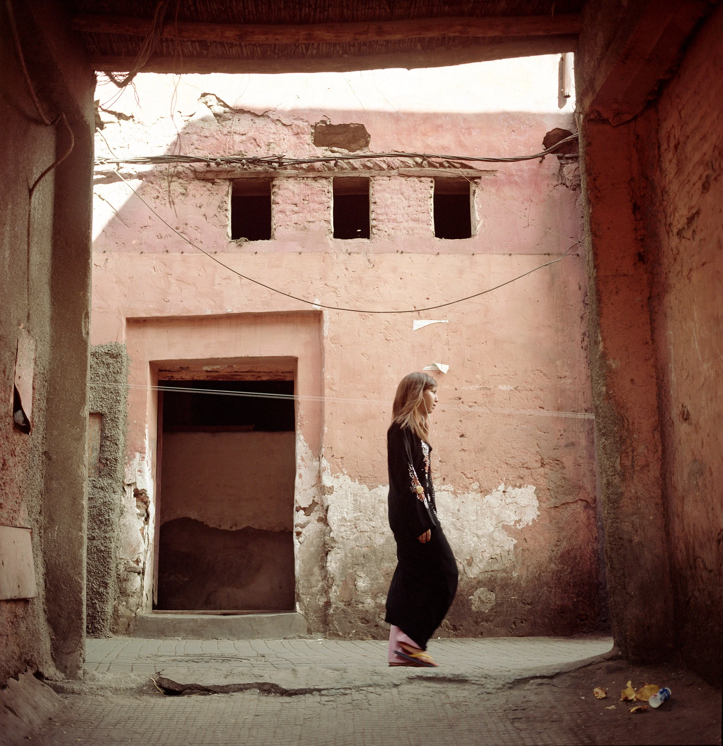 Woman wearing black coat walking in an alley with weathered pink walls and holes in the upper part of a building.