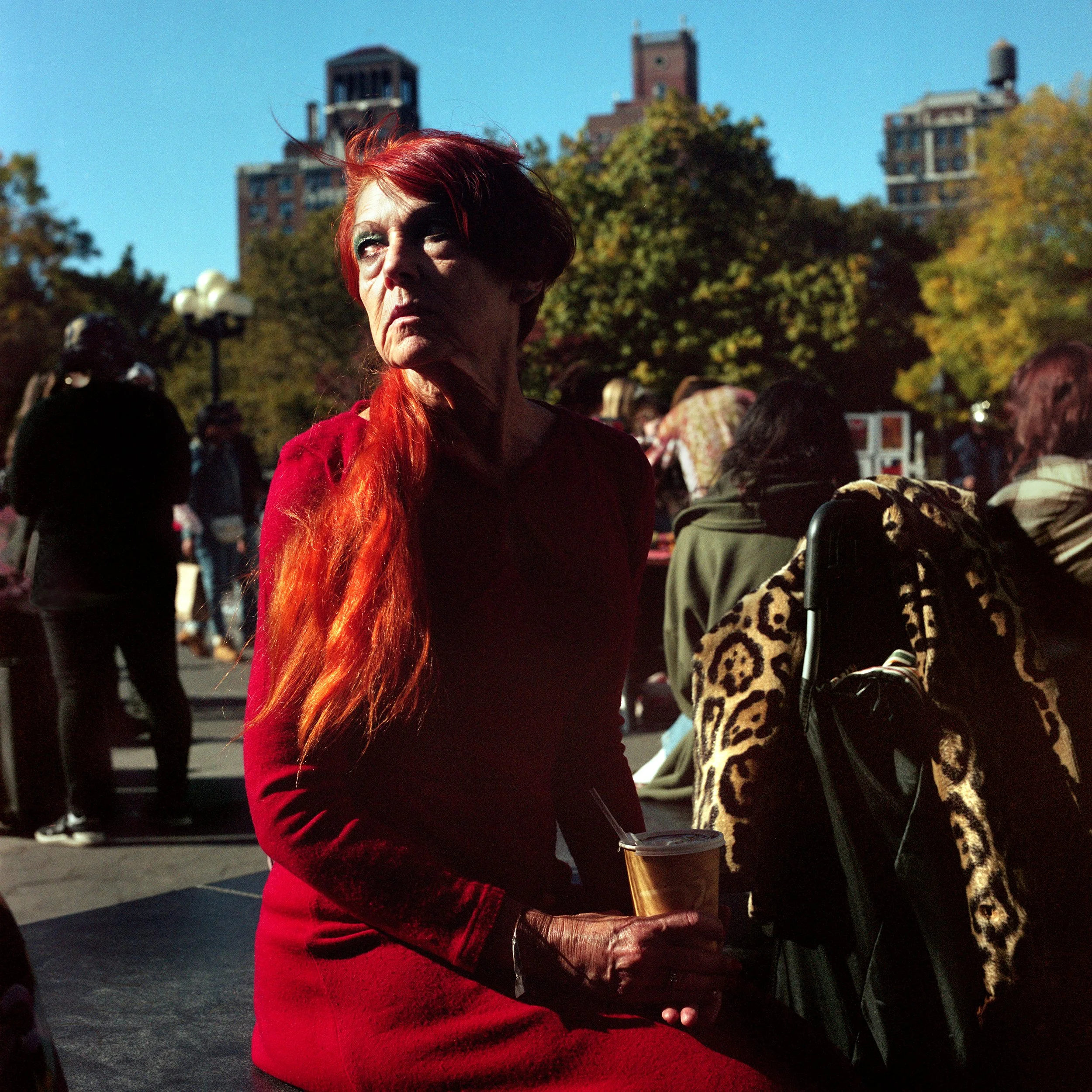 In New York Manhattan, A woman with long red hair wearing a red dress, sitting outdoors among several people in what appears to be a park or public gathering on a sunny day, holding a beverage in her hand.