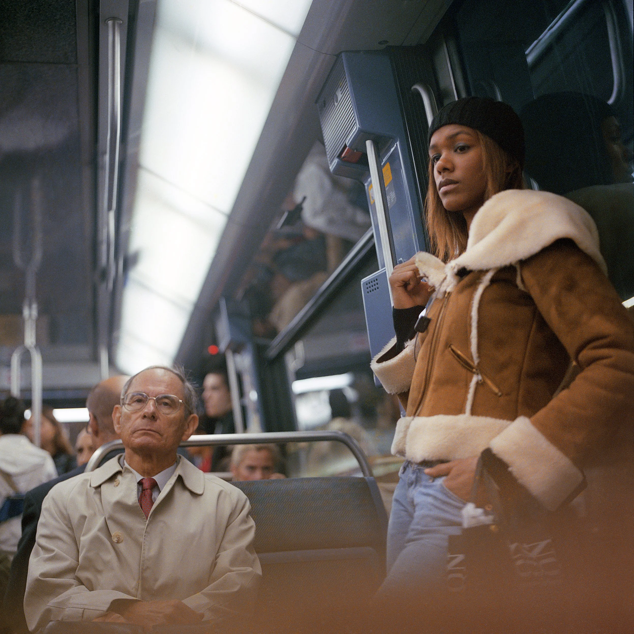 A young woman with long hair wearing a black beanie and a brown shearling jacket stands next to a seated older man wearing glasses and a beige trench coat inside a crowded subway train.
