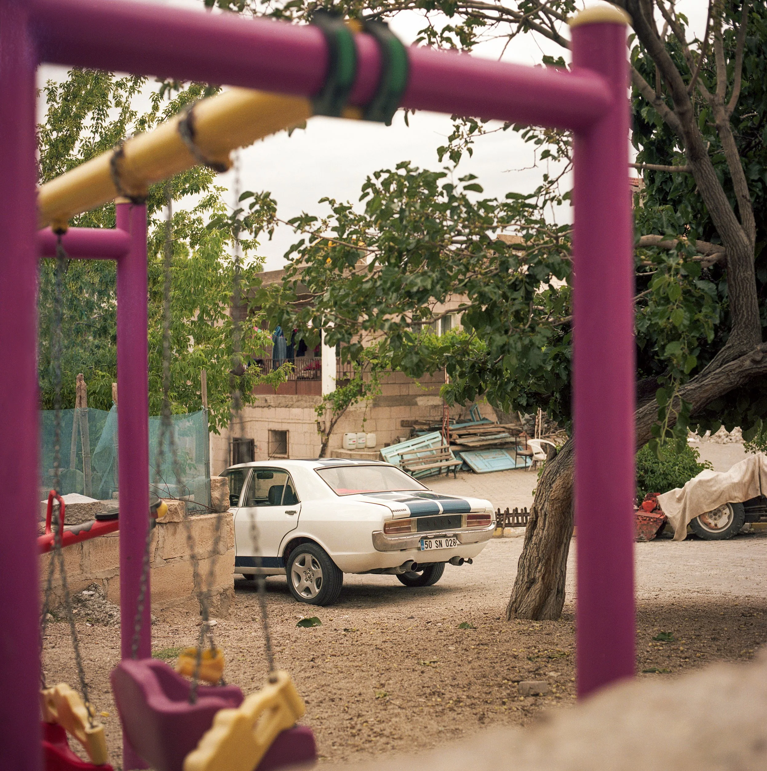 	
In Goreme, Turkey, A white vintage car parked on a dirt ground near a tree and a concrete wall, with playground equipment partially visible in the foreground.