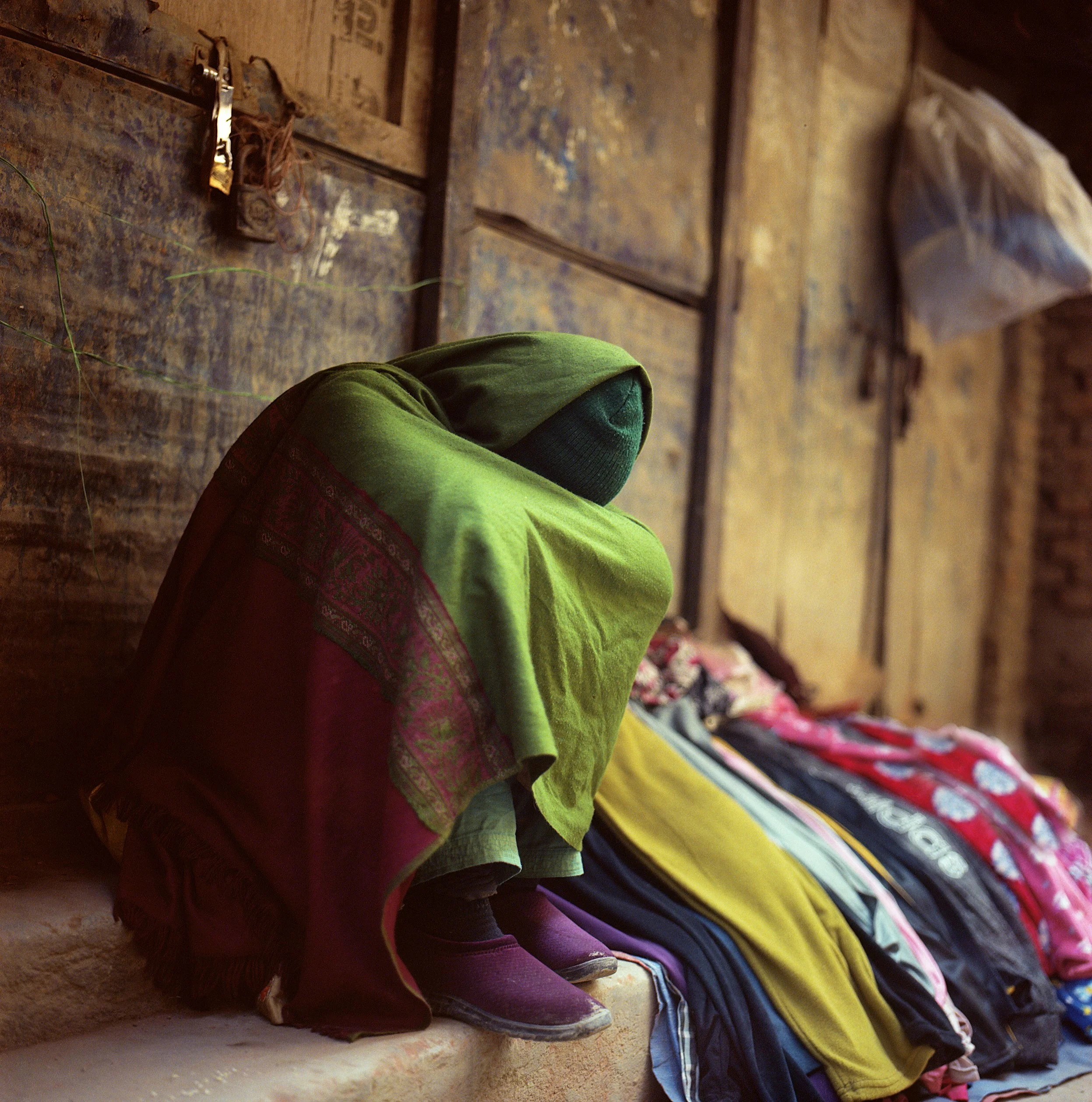 In Kathmandu, Nepal, a person is draped under a green clothe, winter, pavement, A row of colorful children's shoes and boots lined up against a wooden wall, with a focus on the shoes in the foreground and the shoe soles visible.