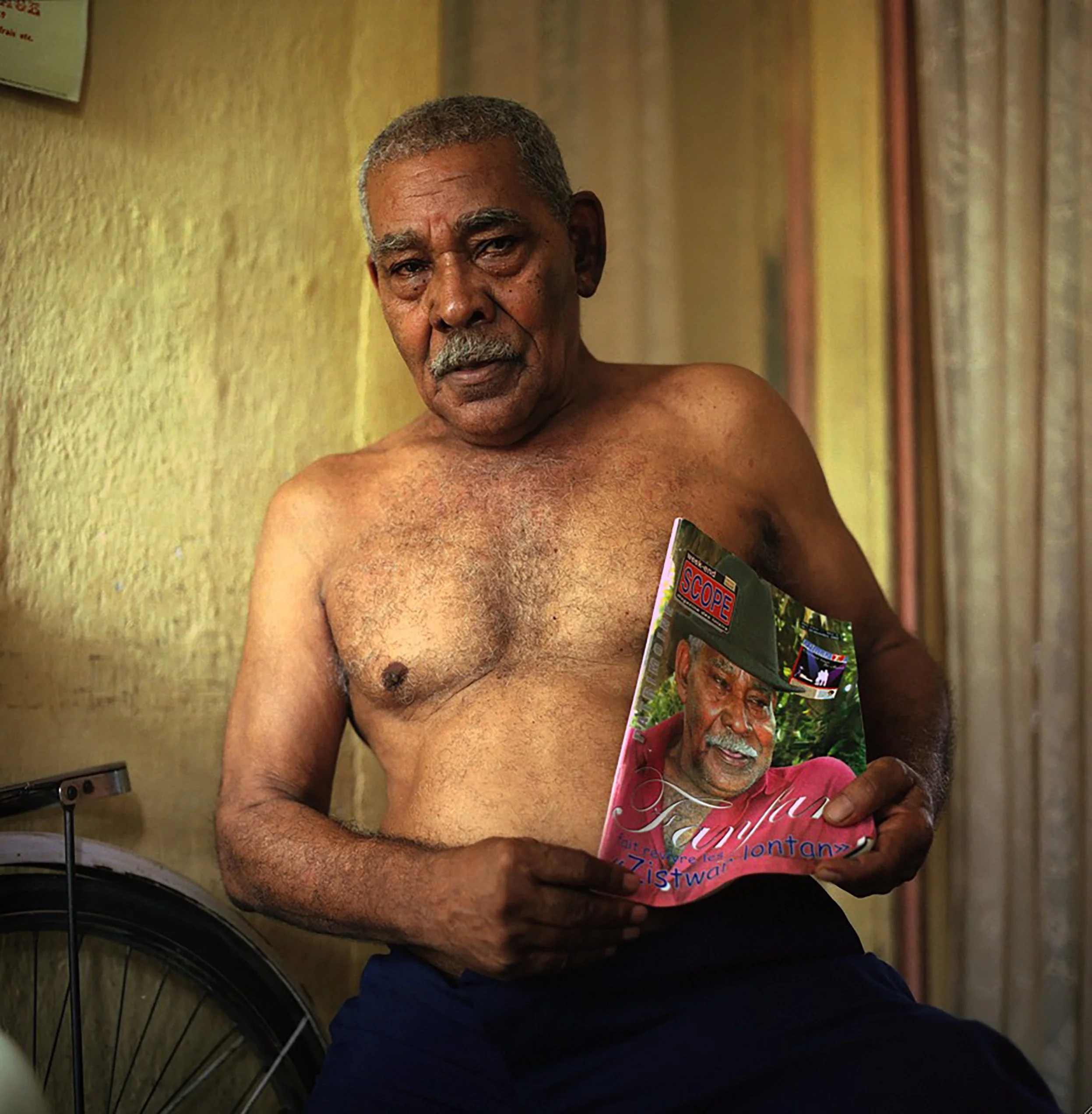 In Mauritius, famous sega singer Fanfan, An elderly man with short gray hair and a mustache sitting shirtless, holding a colorful magazine with his face on the front page, with a yellow wall with curtains in the background.