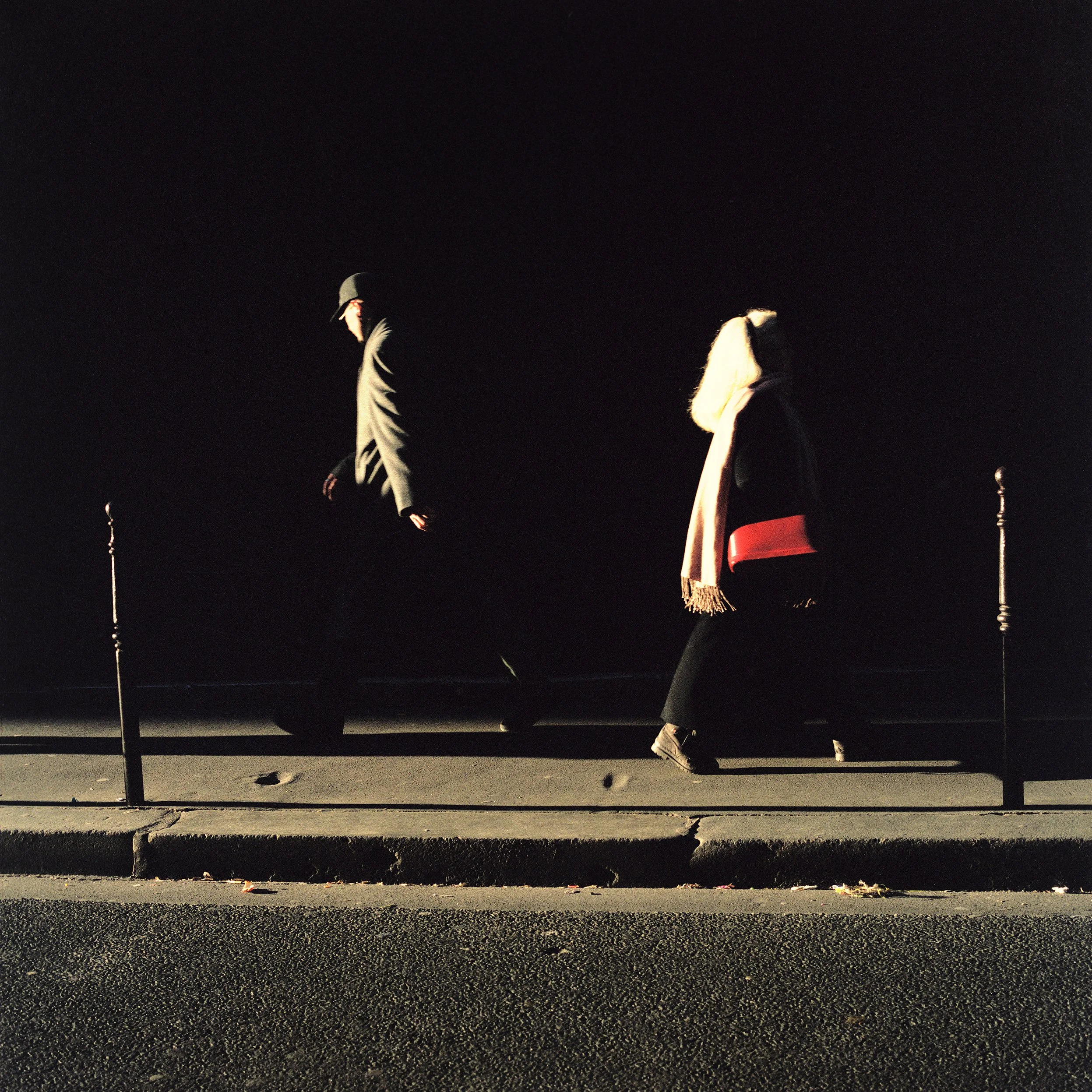 Two people walking on a sidewalk against a dark background, with street poles and shadows visible.