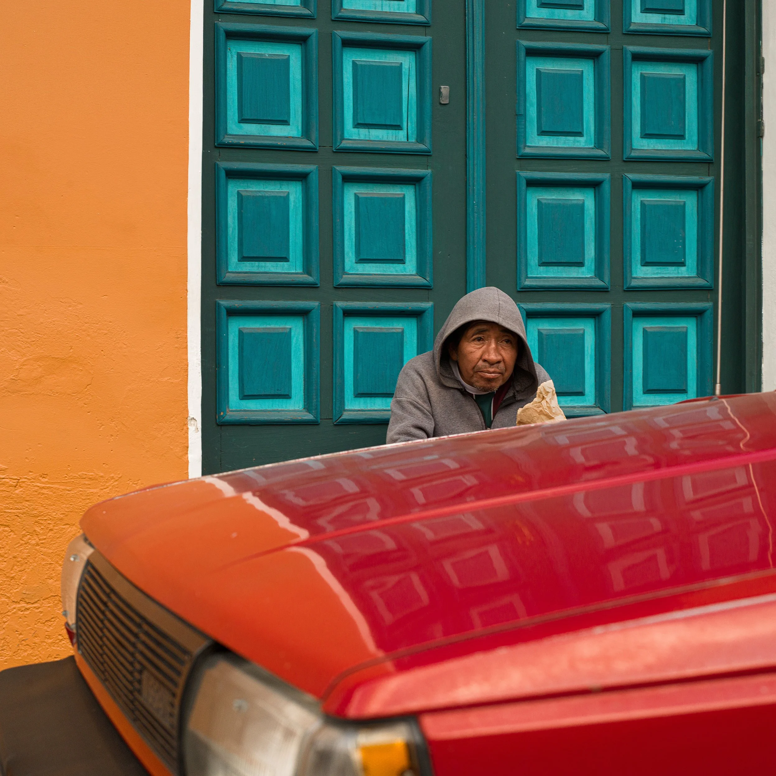 In Antigua, Guatemala, A person in a gray hoodie sitting in front of a turquoise wall with square patterned tiles, a yellow wall border on the left, and part of a red vehicle in the foreground.