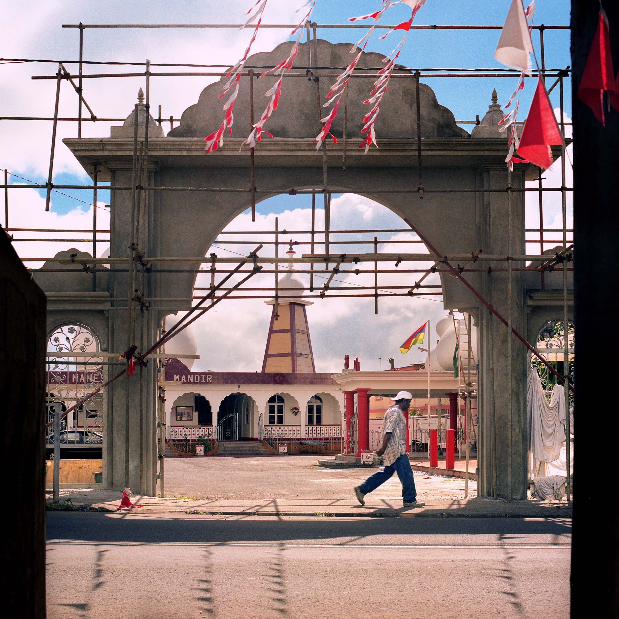 A person walking past a construction archway with scaffolding, in front of a temple-like building with a spire, flags, and decorative architecture.