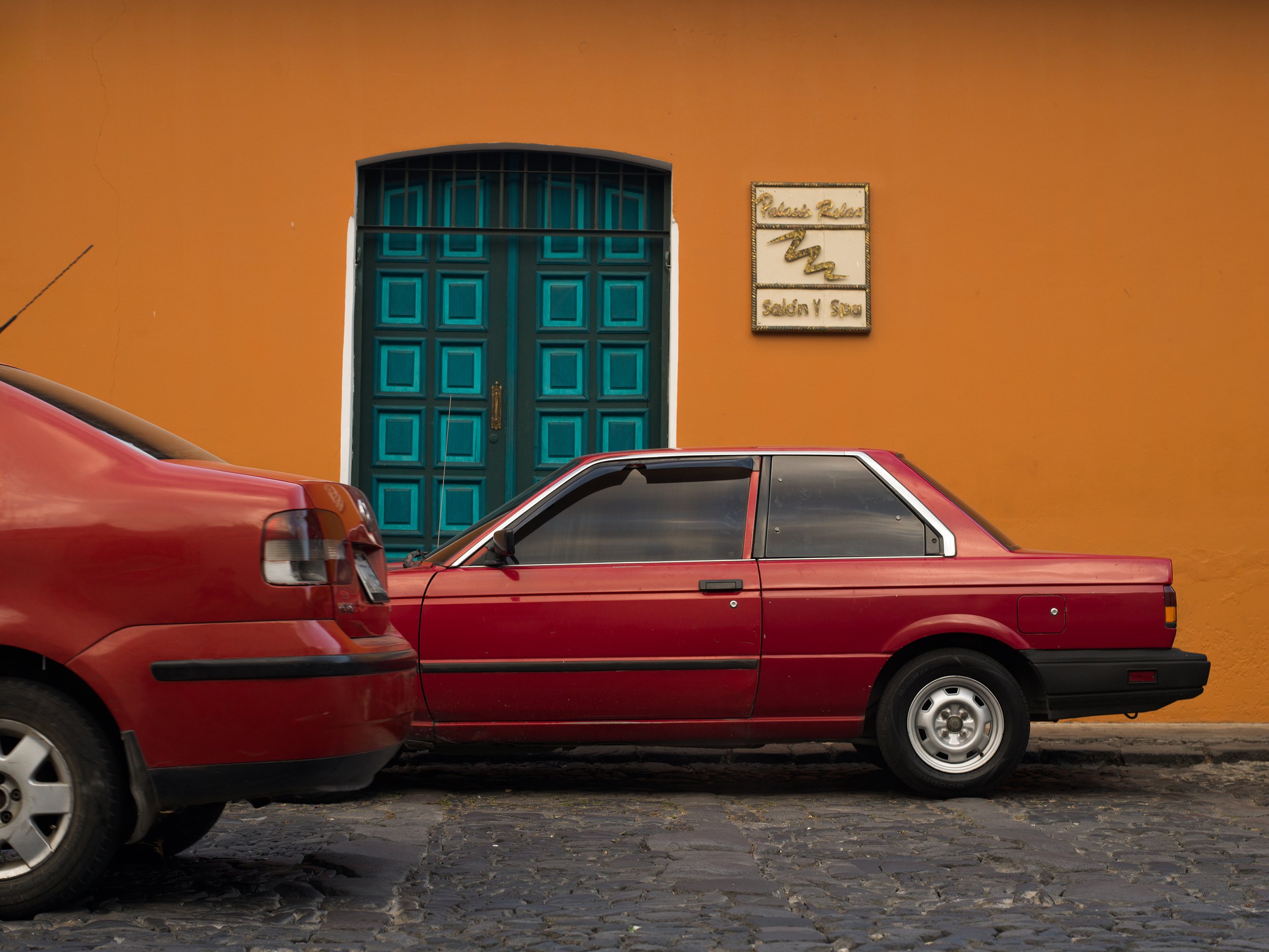 In Antigua, Guatemala, Two red cars parked on a cobblestone street in front of a yellow wall with a teal door and a sign that reads 'Palace Relax Salon & Spa.'