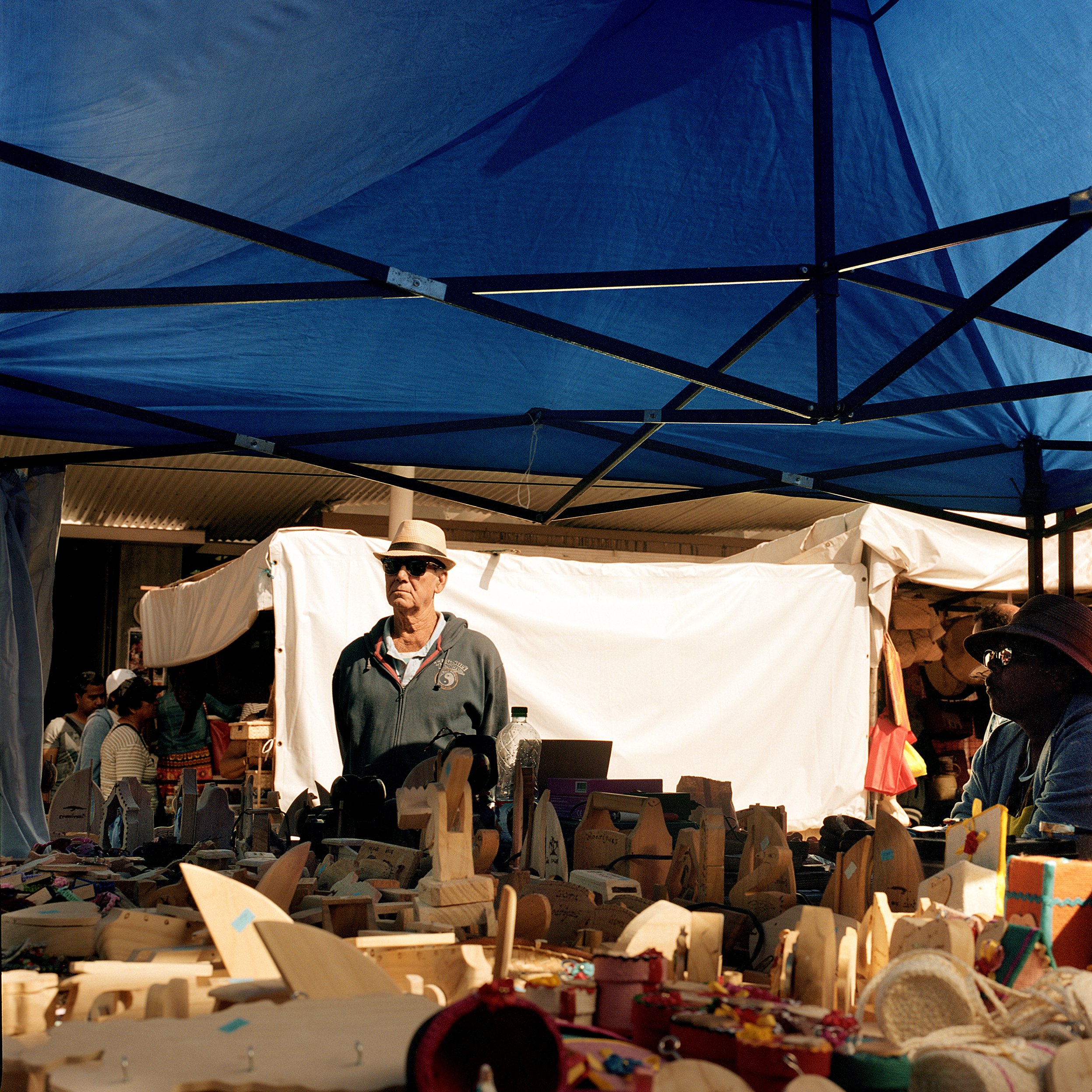 People browsing handmade wooden crafts at an outdoor market stall under a large blue canopy.