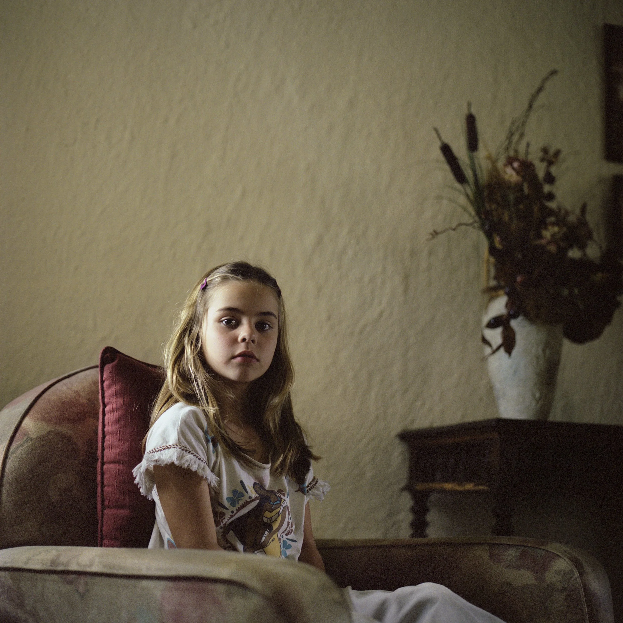 In Mauritius, A young girl with long brown hair, wearing a white shirt with colorful print, sitting on a plush armchair with a maroon cushion, looking directly at the camera.