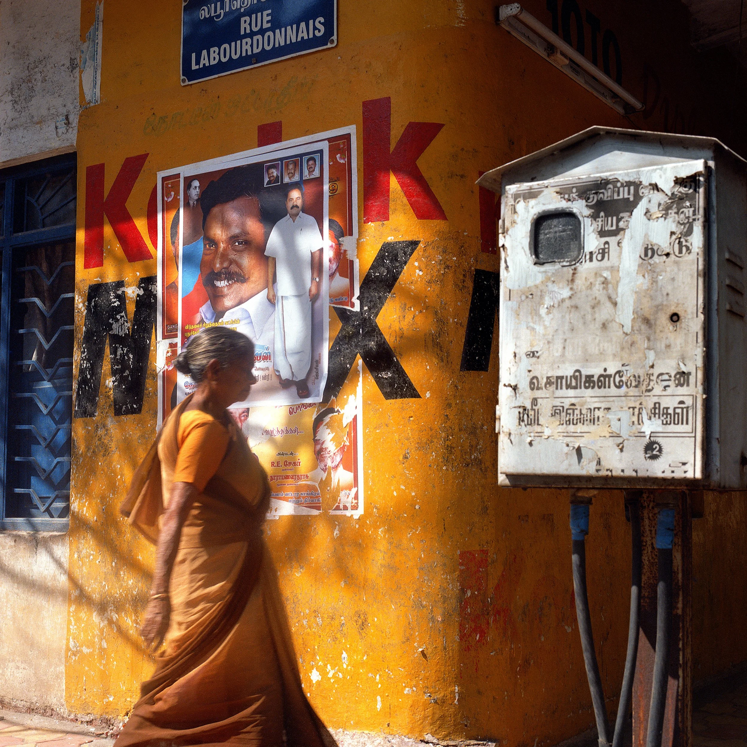 An elderly woman walking past a brightly painted yellow wall with political posters and a blue street sign, near a rusty electrical box.