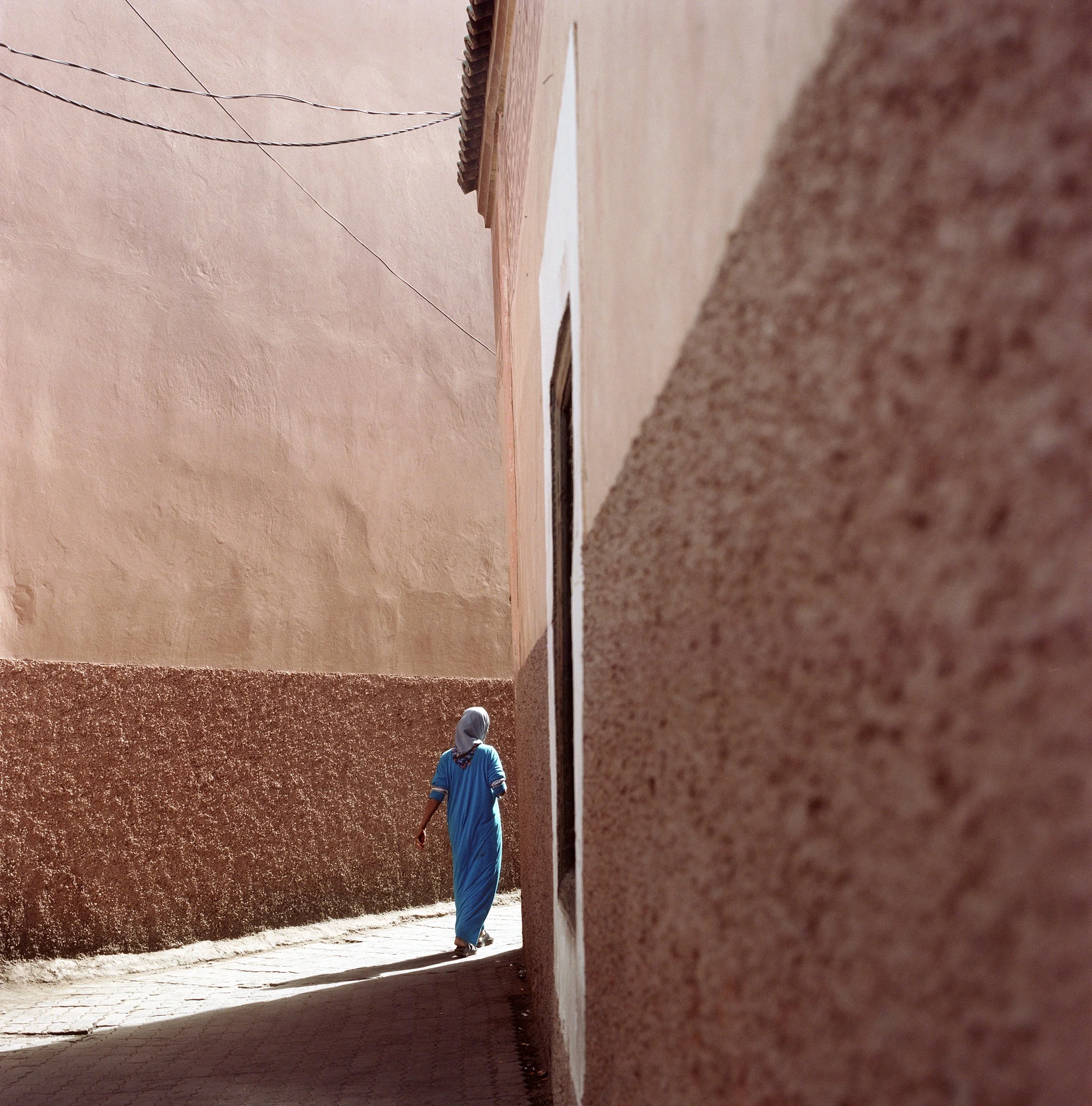 A person wearing a blue garment and headscarf walking down a narrow alleyway with textured walls on either side.