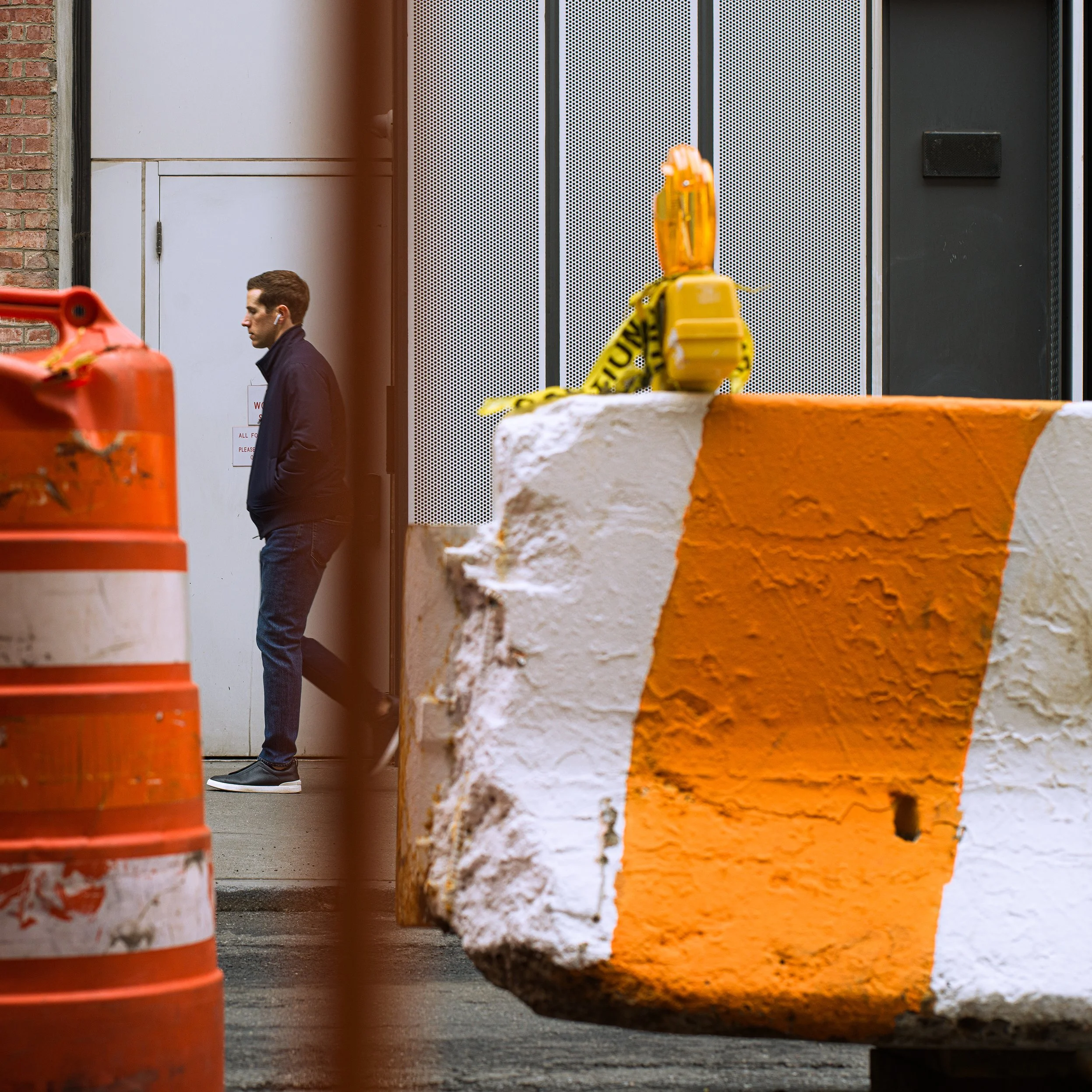 In New York Manhattan, A man standing on a city sidewalk near orange and white construction barriers and pipes, with a brick and modern building facade in the background.